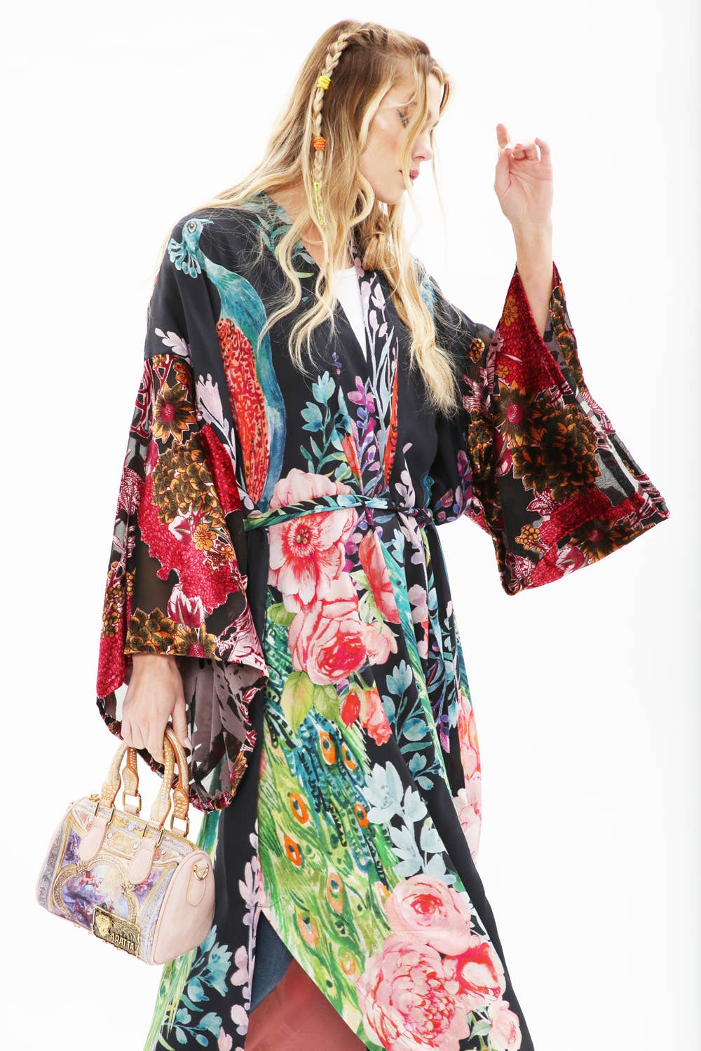 A woman with long blonde hair models the Sweet Fantasy Kimono by Aratta—a peacock floral dress with wide burnout velvet sleeves—holding an ornate handbag, standing against a white background and looking to the side.