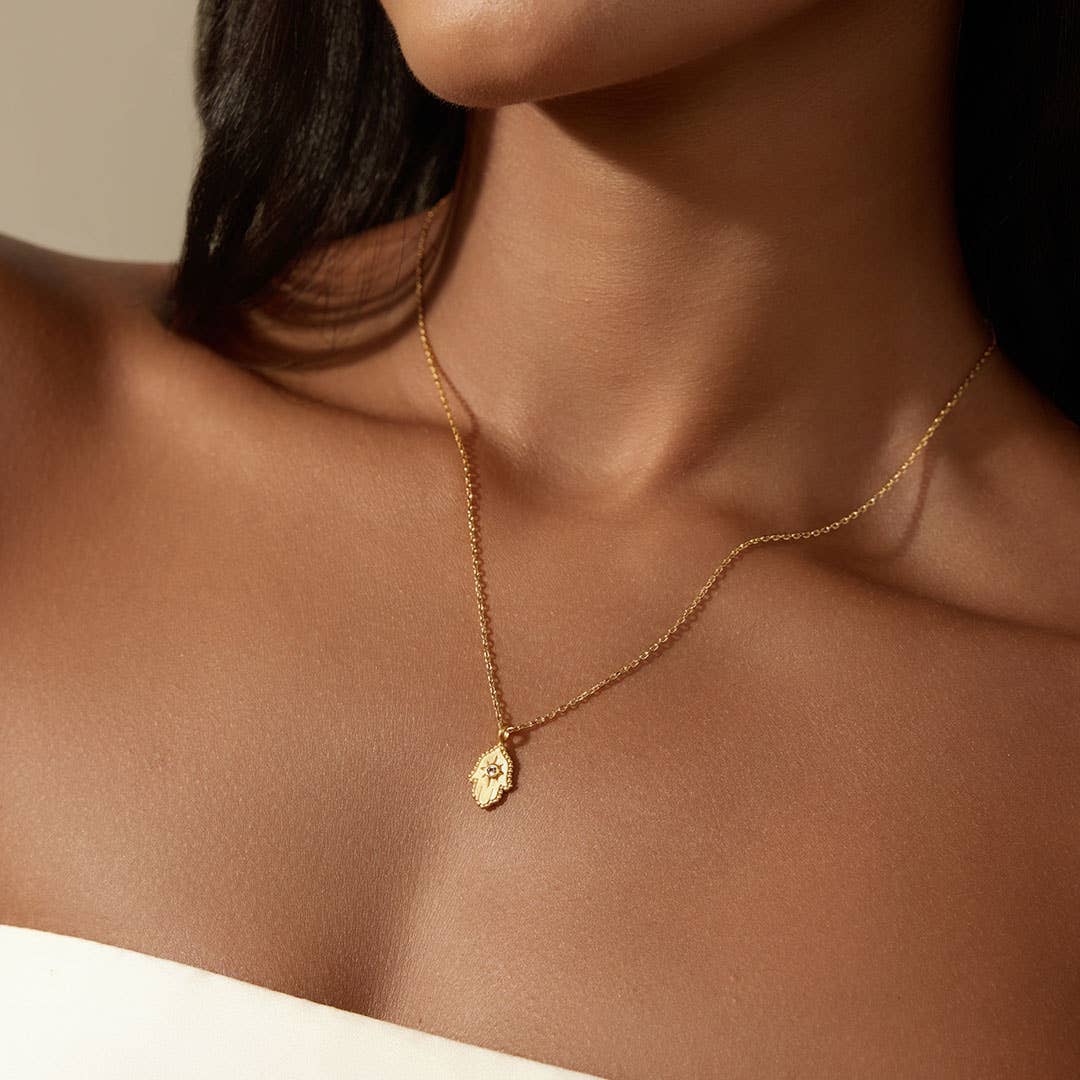 A woman with medium brown skin and straight dark hair wears Satya Jewelry’s Protective Hand White Topaz Hamsa Necklace 16". She’s shown from the shoulders up, dressed in a white strapless top.
