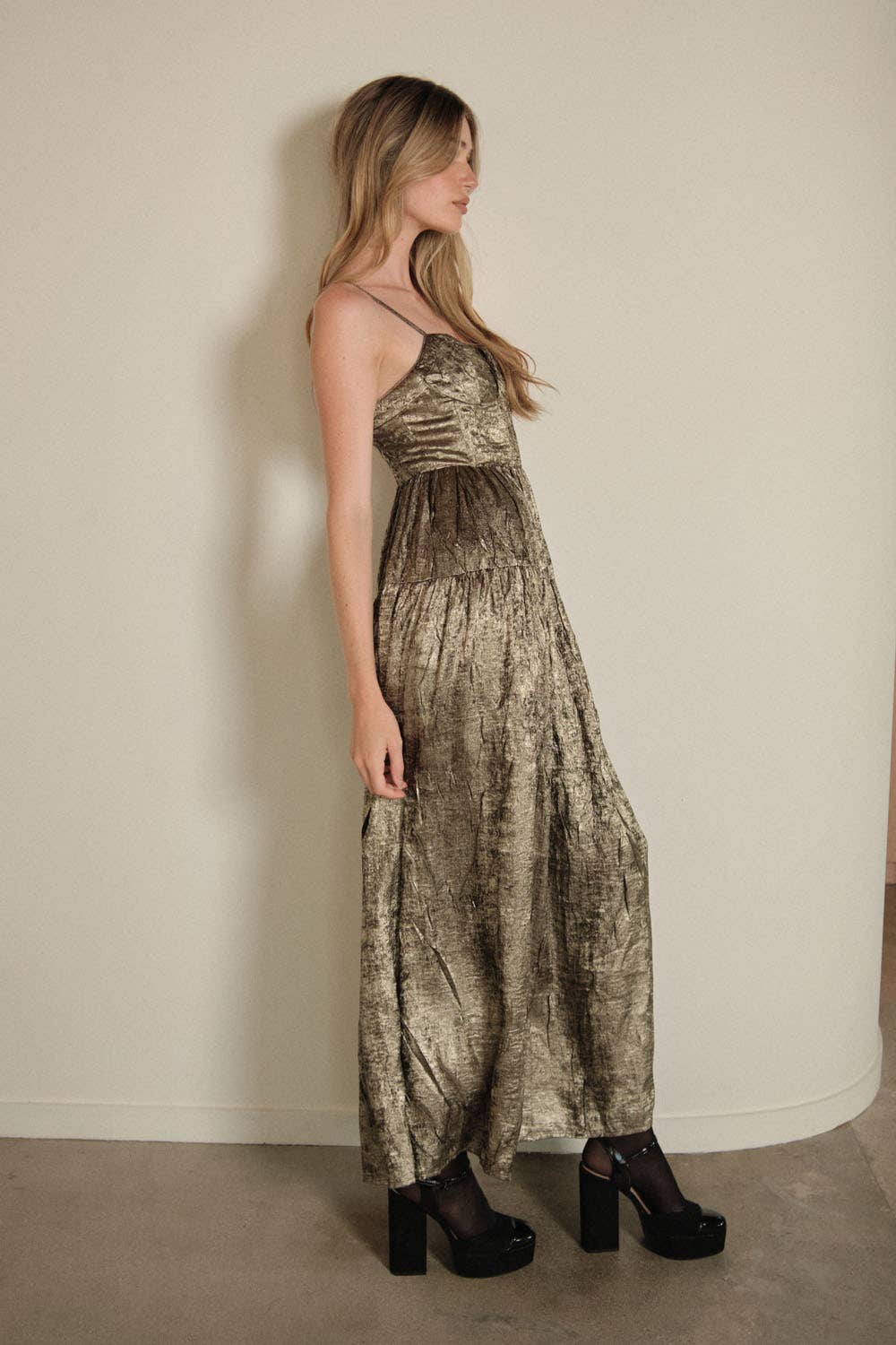 A woman with long, wavy hair faces sideways against a plain wall, wearing the Promesa USA Textured Metallic Fit-and-Flare Corset Maxi Dress and black platform heels in a relaxed pose.