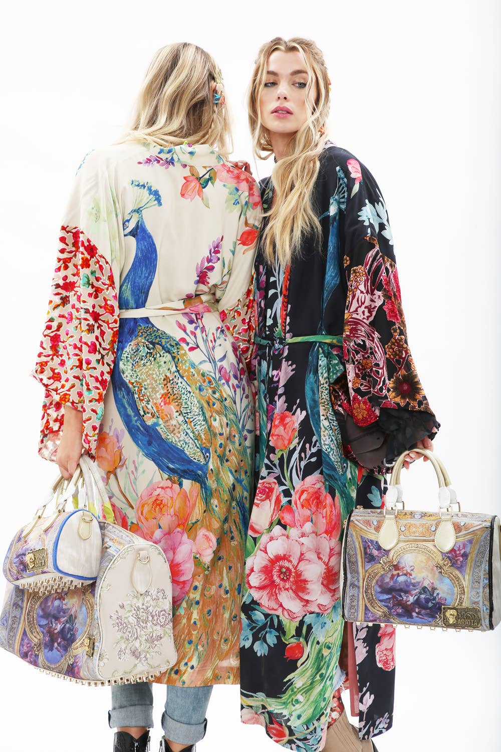 Two women stand side by side in Aratta’s Sweet Fantasy Kimonos, featuring vibrant peacock florals and burnout velvet sleeves. Each holds an ornate handbag, their wavy hair and the white background highlighting the bold kimono designs.