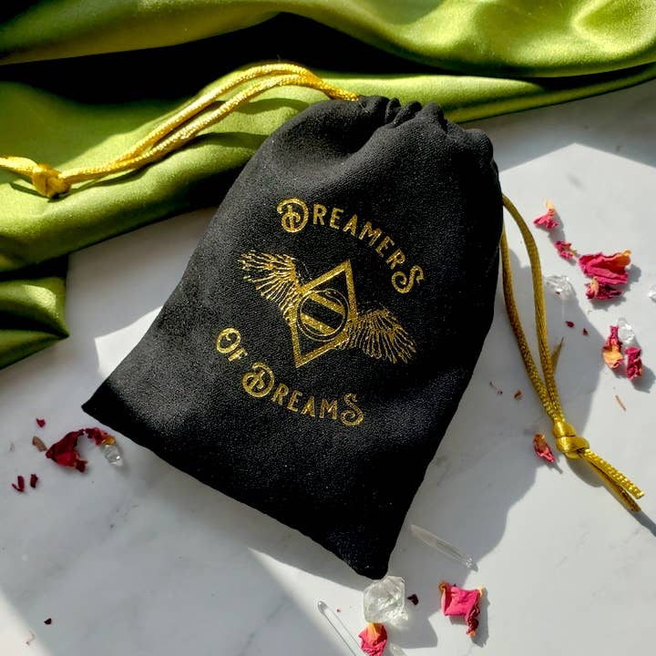A black drawstring pouch with gold cords and Dreamers of Dreams in gold sits on a white surface with rose petals, crystals, and the Dreamers of Dreams Big Gold Heart | Paperclip Chain Necklace beside green fabric.