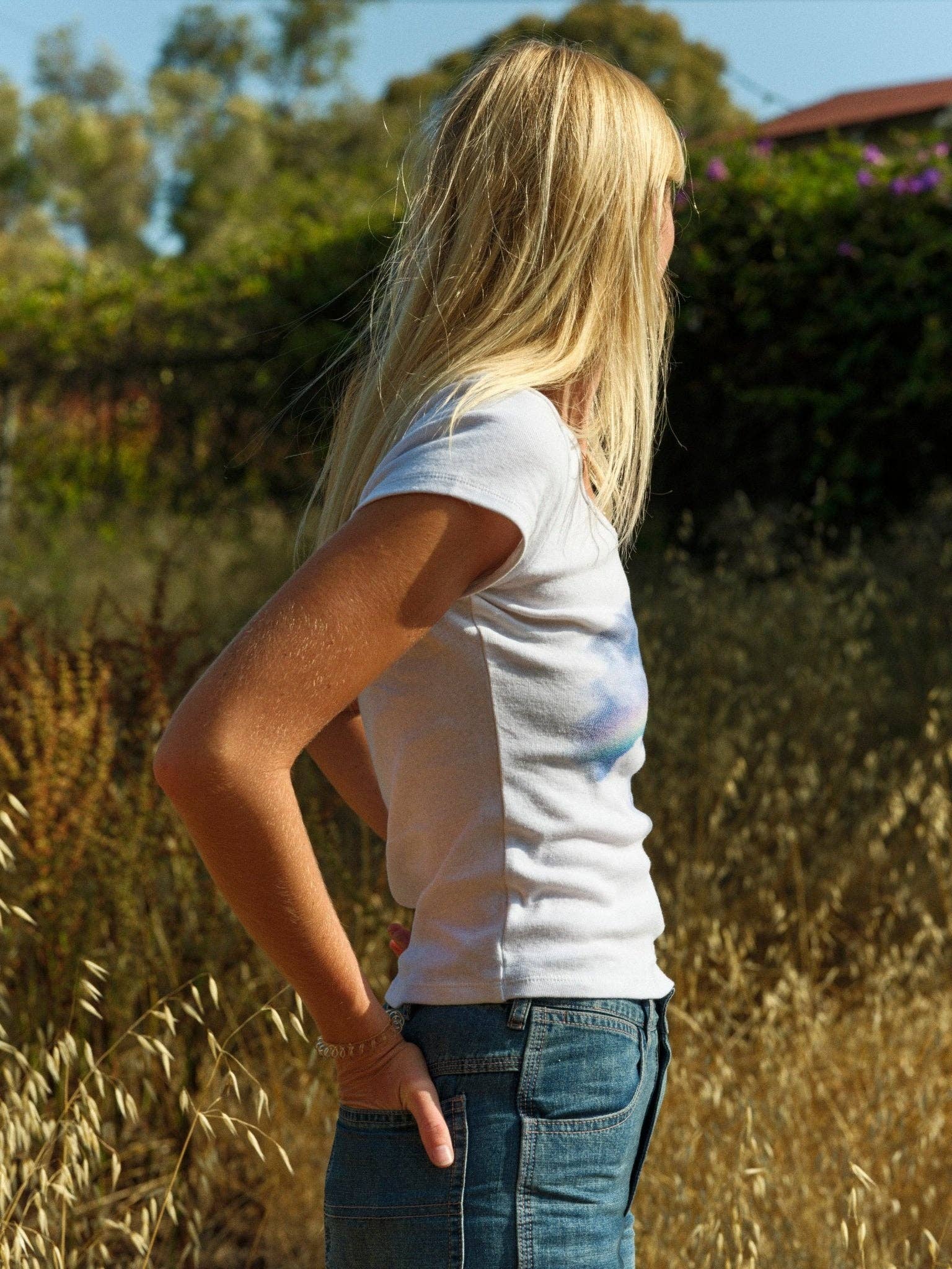 A person with long blonde hair stands outdoors in a sunlit grassy area, wearing blue jeans and the Mollusk CloudBreak Tee, inspired by California surf culture, with shrubs and trees in the background.
