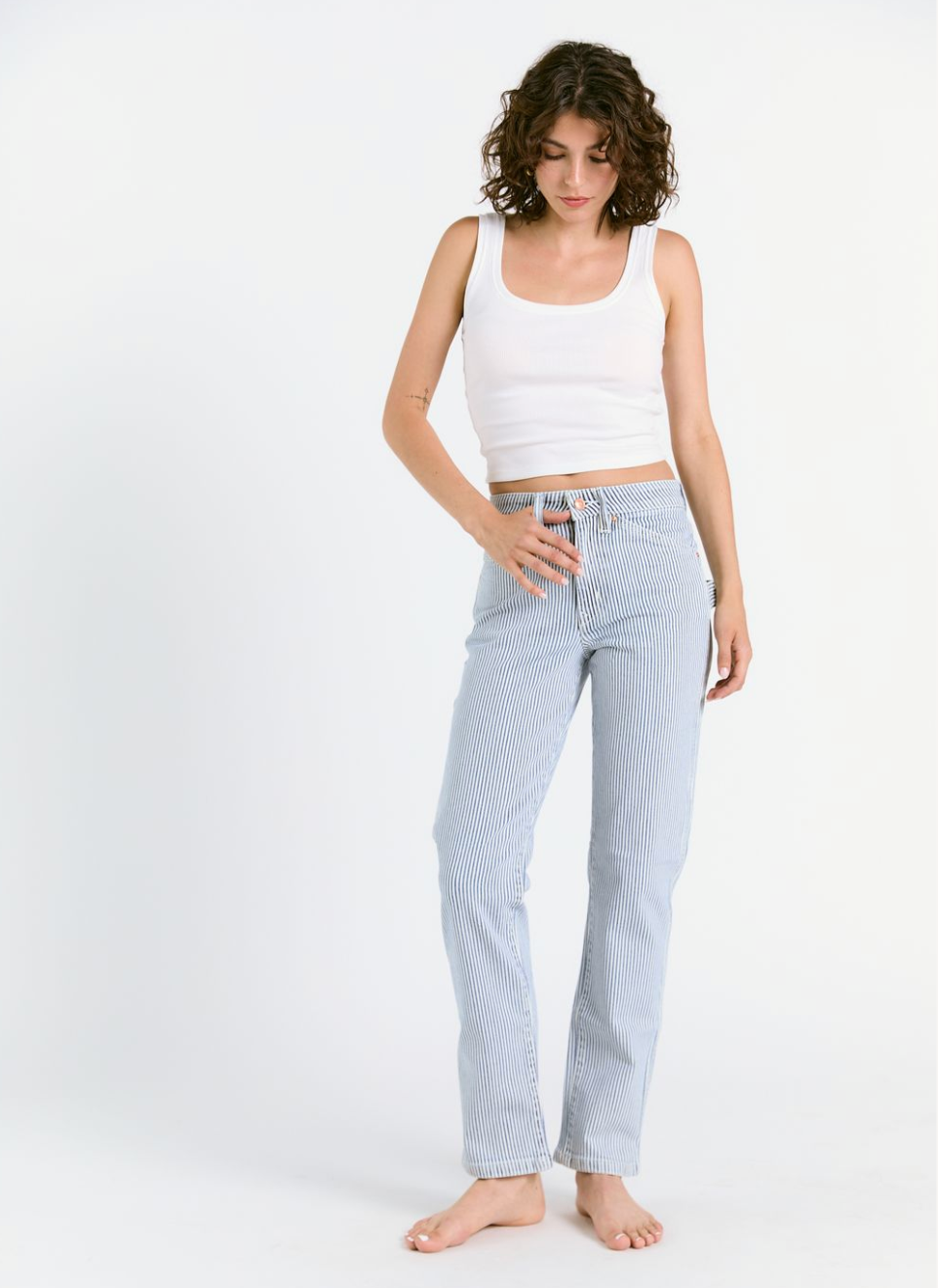 A woman with short curly hair models Porter Blue Apparel’s PEYTON PAINTER IN PACIFIC WASH, pairing the relaxed straight carpenter pants with a white tank top. She stands barefoot against a plain white background, looking down with one hand on her hip.