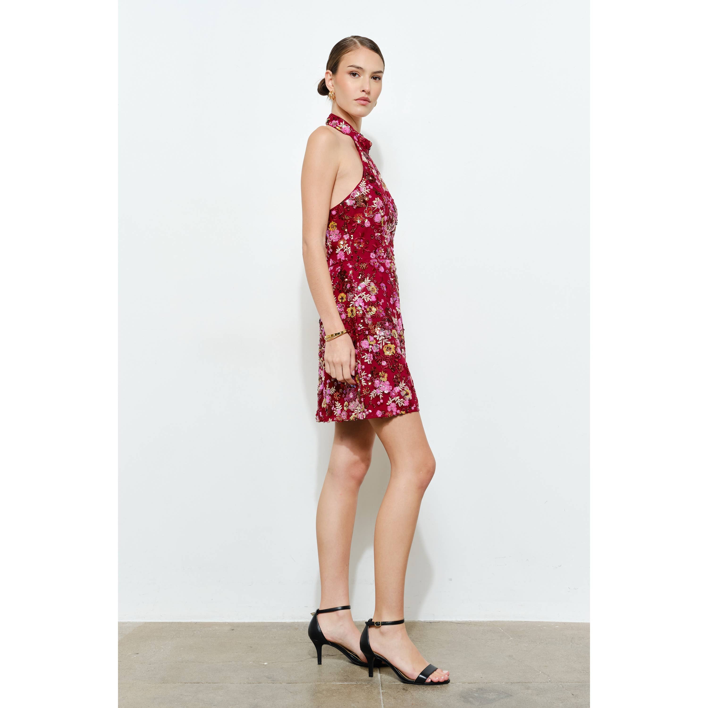 Model wearing a red floral sequins embellished halter neck mini dress with black ankle-strap heels against white wall