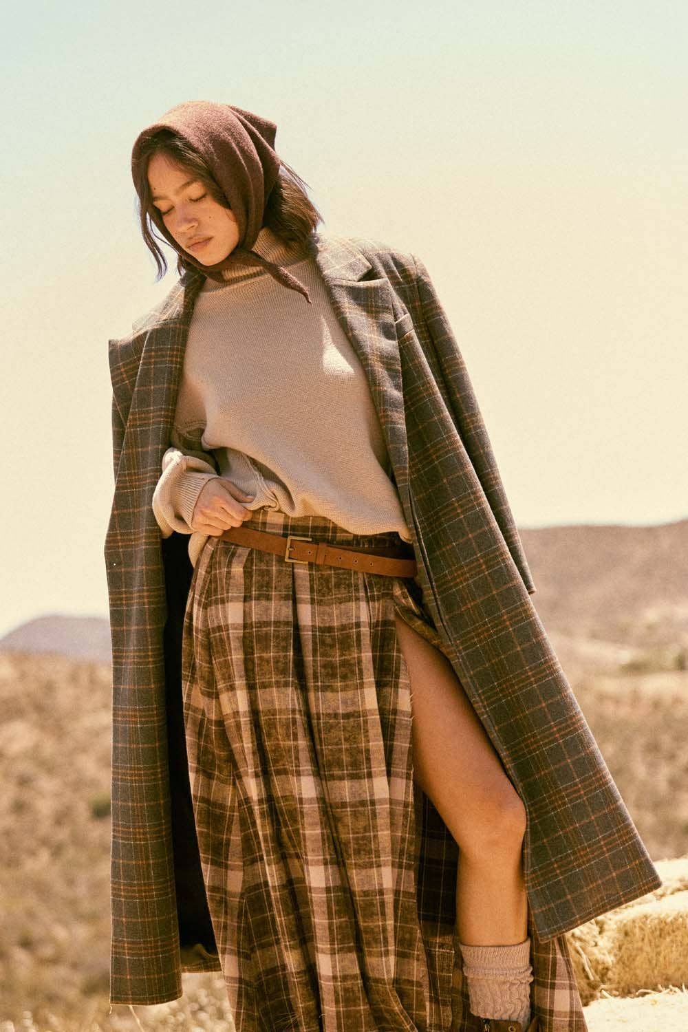 A woman stands outdoors in a Promesa USA Plaid Felt Double-Breasted Calf-Length Overcoat, paired with a brown headscarf, beige turtleneck sweater, plaid skirt with slit, brown belt, and gray leg warmers against a dry, hilly landscape.