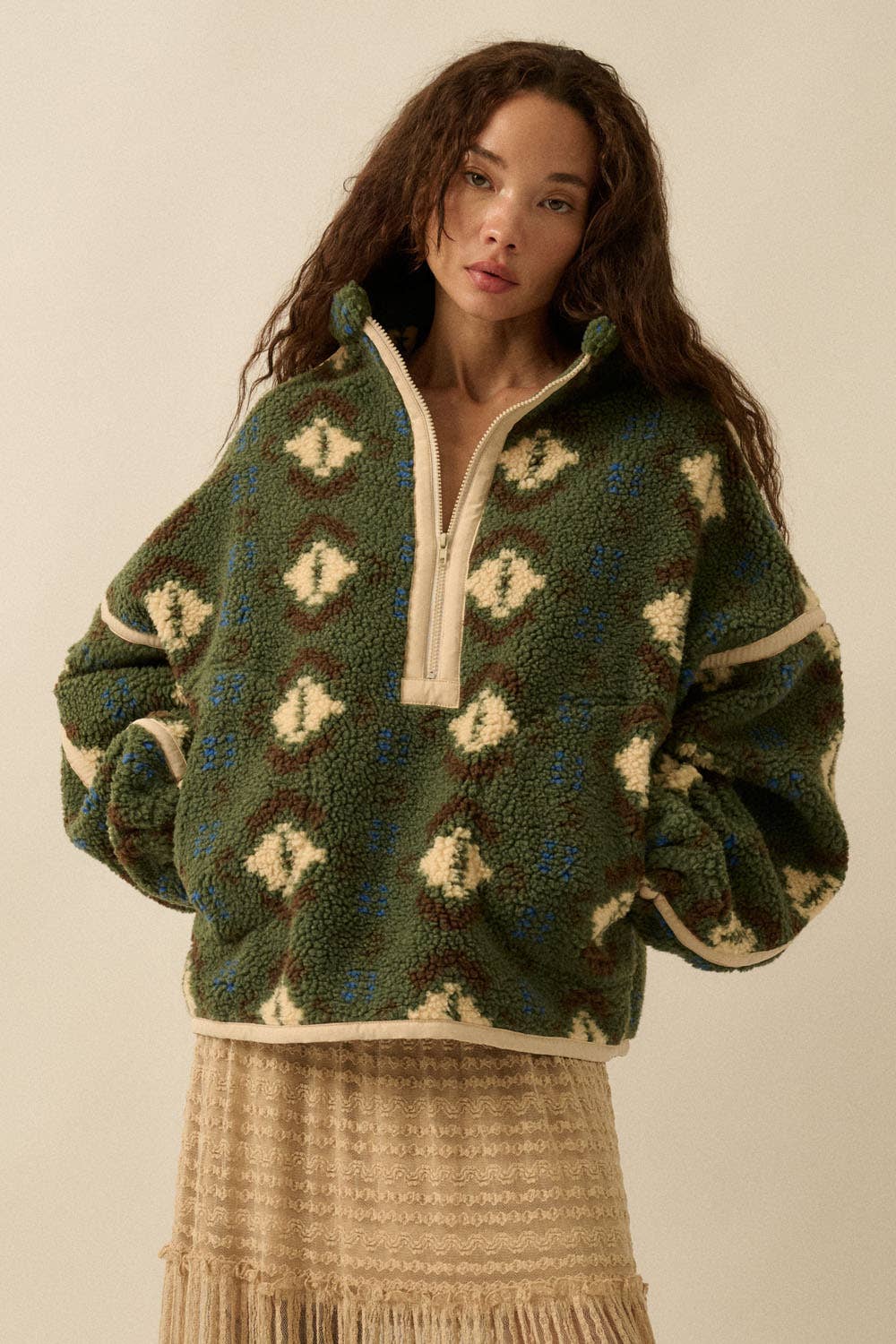 A woman with long, wavy brown hair models the Promesa USA Geo-Print Sherpa Fleece Half-Zip Pullover Jacket, styled with a beige fringe-layered skirt, against a plain light background.