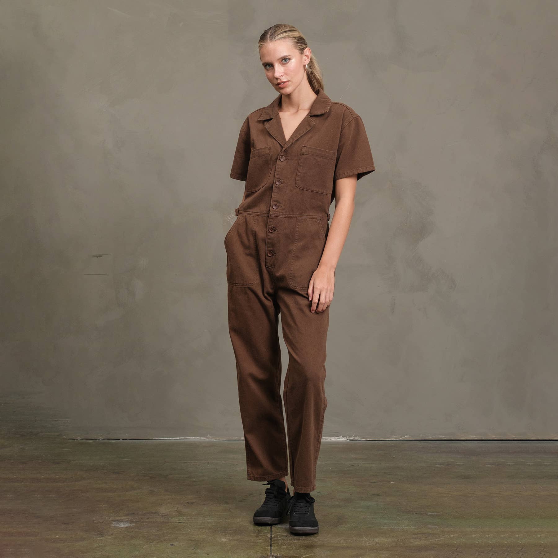 A woman stands indoors against a textured gray wall, wearing the RAISONNEL Field Jumpsuit - RJP1001 in brown cotton with black shoes. Hands in her pockets, she gazes slightly to the side.