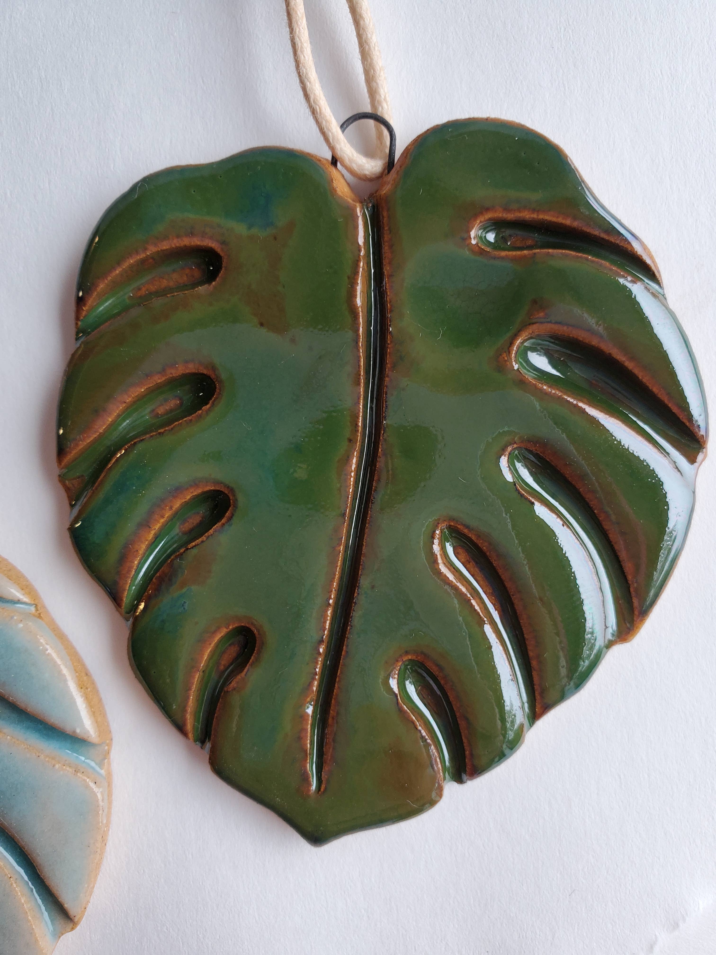 Glossy green ceramic Monstera leaf ornament with textured cutouts and hanging loop on a light background.