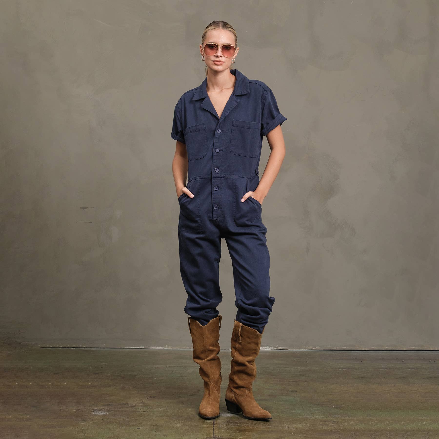 A person wearing sunglasses, the RAISONNEL Field Jumpsuit - RJP1001 with rolled sleeves and cuffs, and brown suede knee-high boots stands against a plain gray wall with hands in pockets.