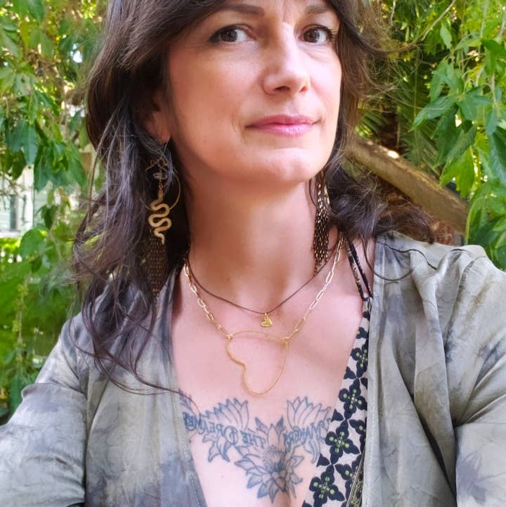 A woman with long brown hair and earrings poses outdoors before green foliage, wearing the Big Gold Heart Paperclip Chain Necklace by Dreamers of Dreams, a patterned top, and displaying a large floral chest tattoo.