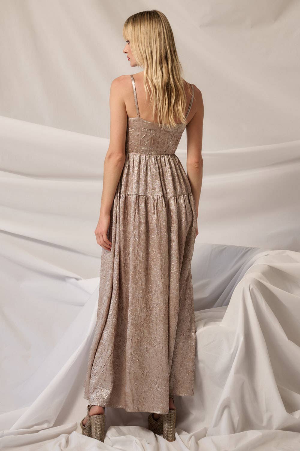A woman with long blonde hair stands with her back to the camera, wearing the Promesa USA Textured Metallic Fit-and-Flare Corset Maxi Dress and platform shoes against a backdrop of light, neutral draped fabric.