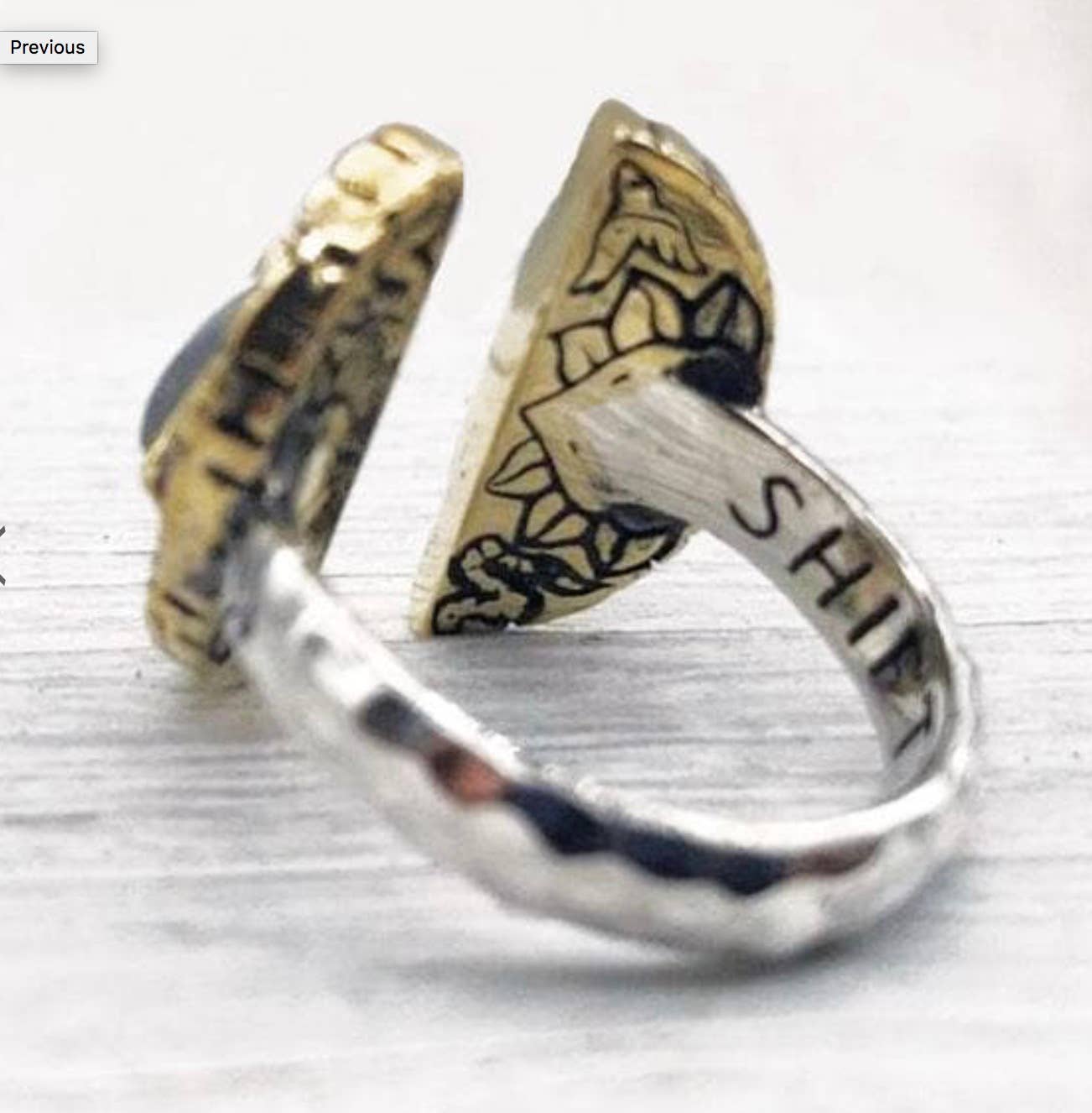 A close-up of the bahgsu jewels Shape Shift Ring in silver and gold, featuring “SHIFT” and floral engravings on the inner band and a dark gemstone accent, showcased as a unique adjustable talisman ring on a light surface.