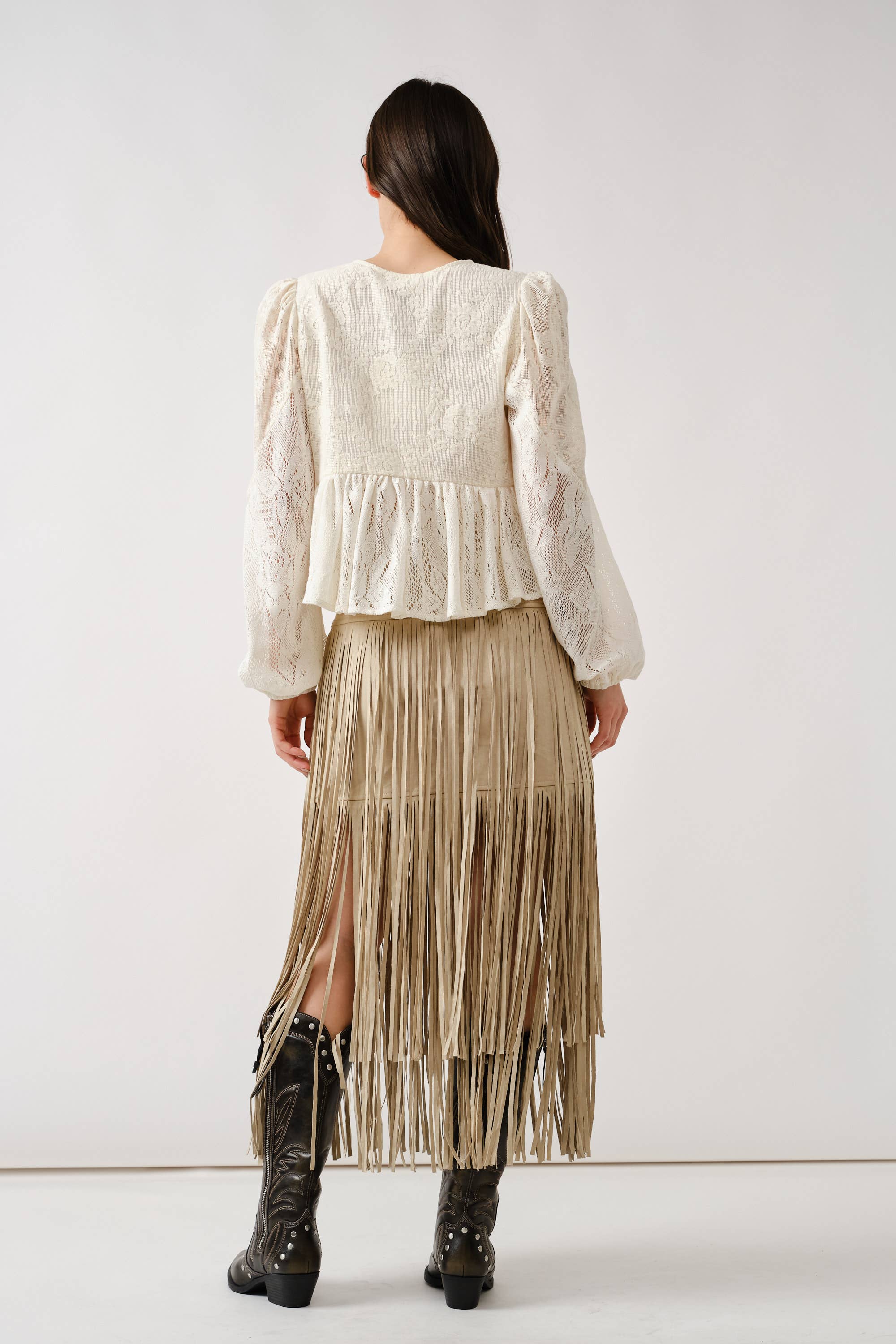A woman with long dark hair faces away, wearing the Aaron & Amber Long Sleeve Lace Babydoll top in cream, styled with a tan fringed suede skirt and black western boots. The background is plain white.
