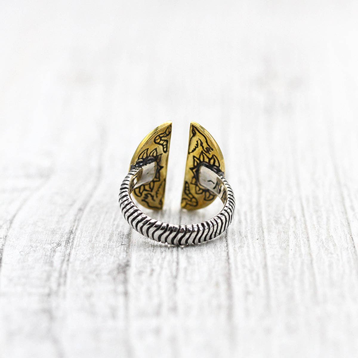 The bahgsu jewels Natural Mystic Ring - Turquoise features a coiled sterling silver band with two open ends, each adorned with golden semi-circular accents and detailed black patterns, displayed on a light wooden surface.