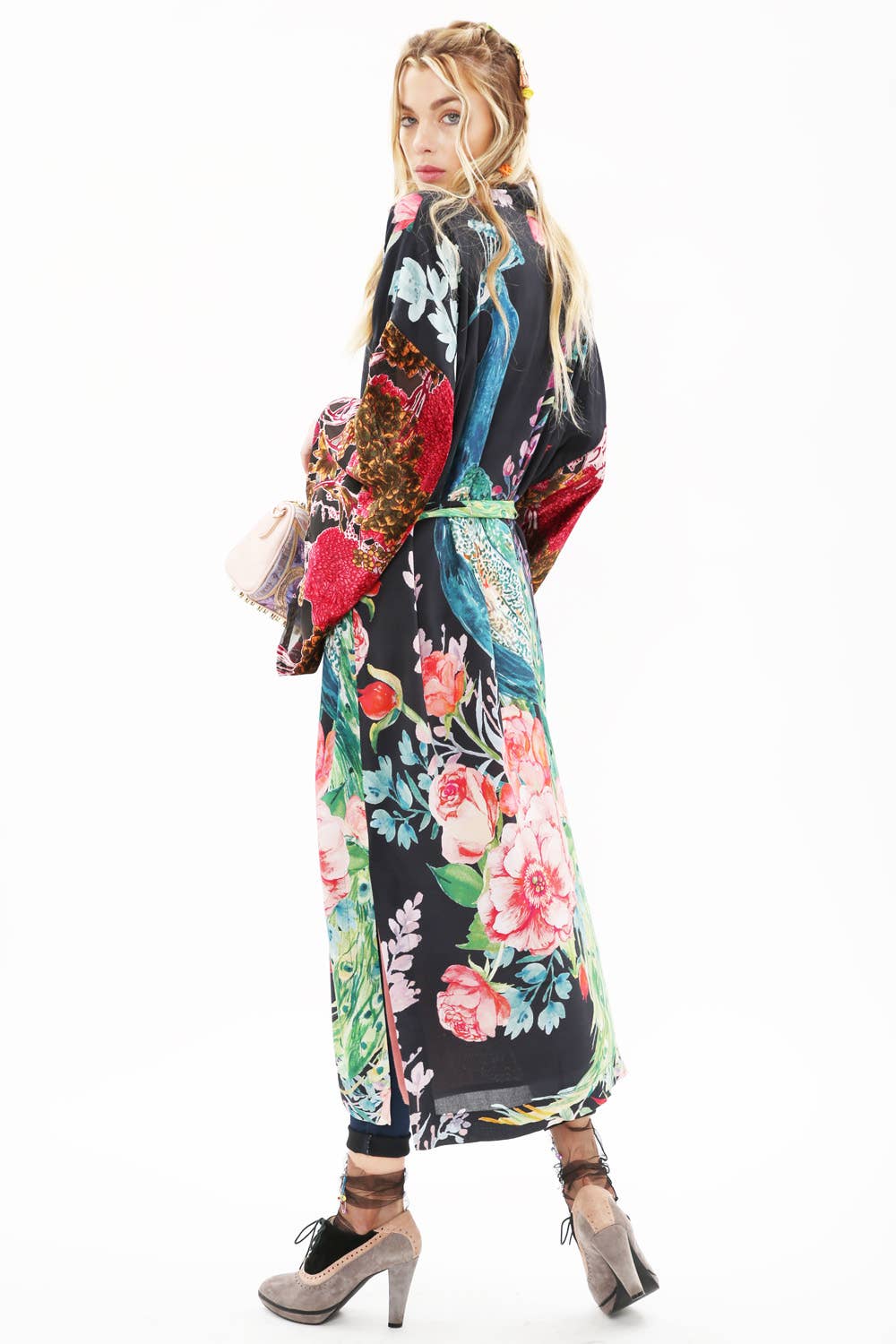 A woman with long blonde hair stands backward, glancing over her shoulder. She wears the Sweet Fantasy Kimono by Aratta with burnout velvet sleeves, paired with dark pants and taupe heels, holding a light pink purse against a white background.