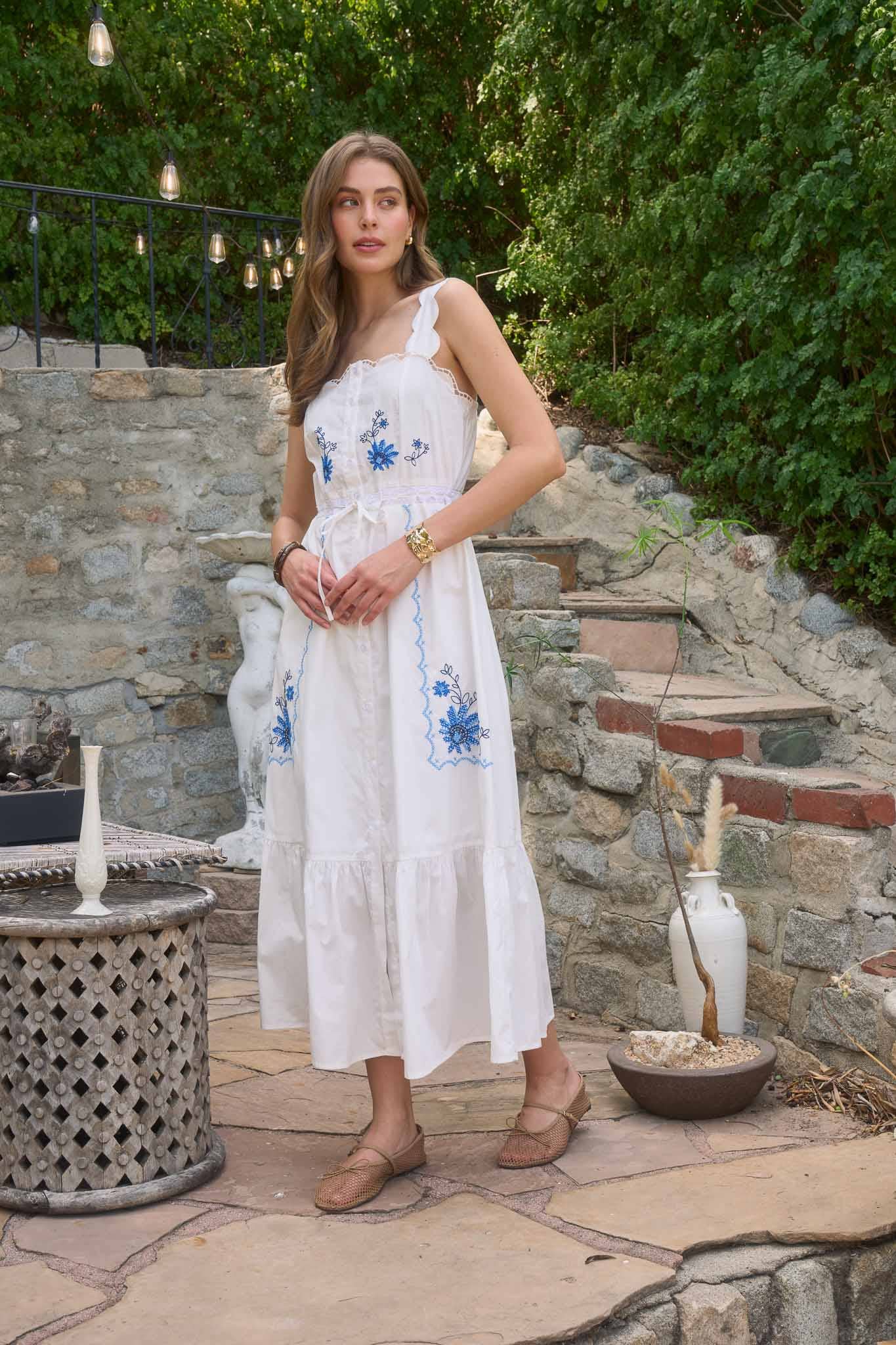 Woman wearing The Molly Embroidered Detail Midi Dress in white with blue floral embroidery in a rustic outdoor setting