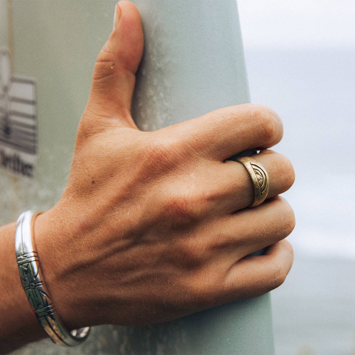 Close-up of a hand wearing Medicine Wheel Cuff || Men in brass, holding a surfboard with ocean in the background