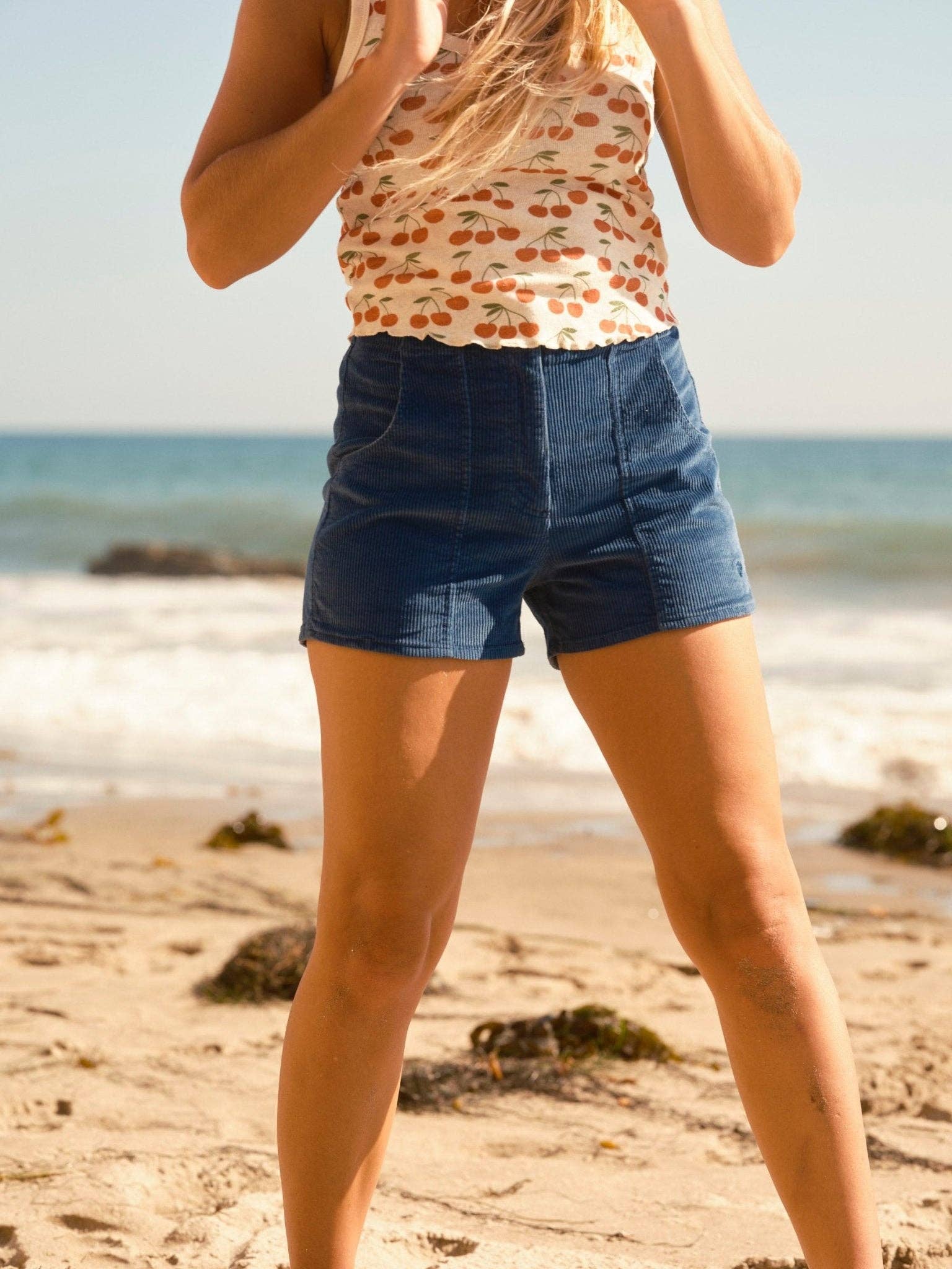 Woman wearing blue Shroom Shorts standing on sunny beach with ocean waves in background
