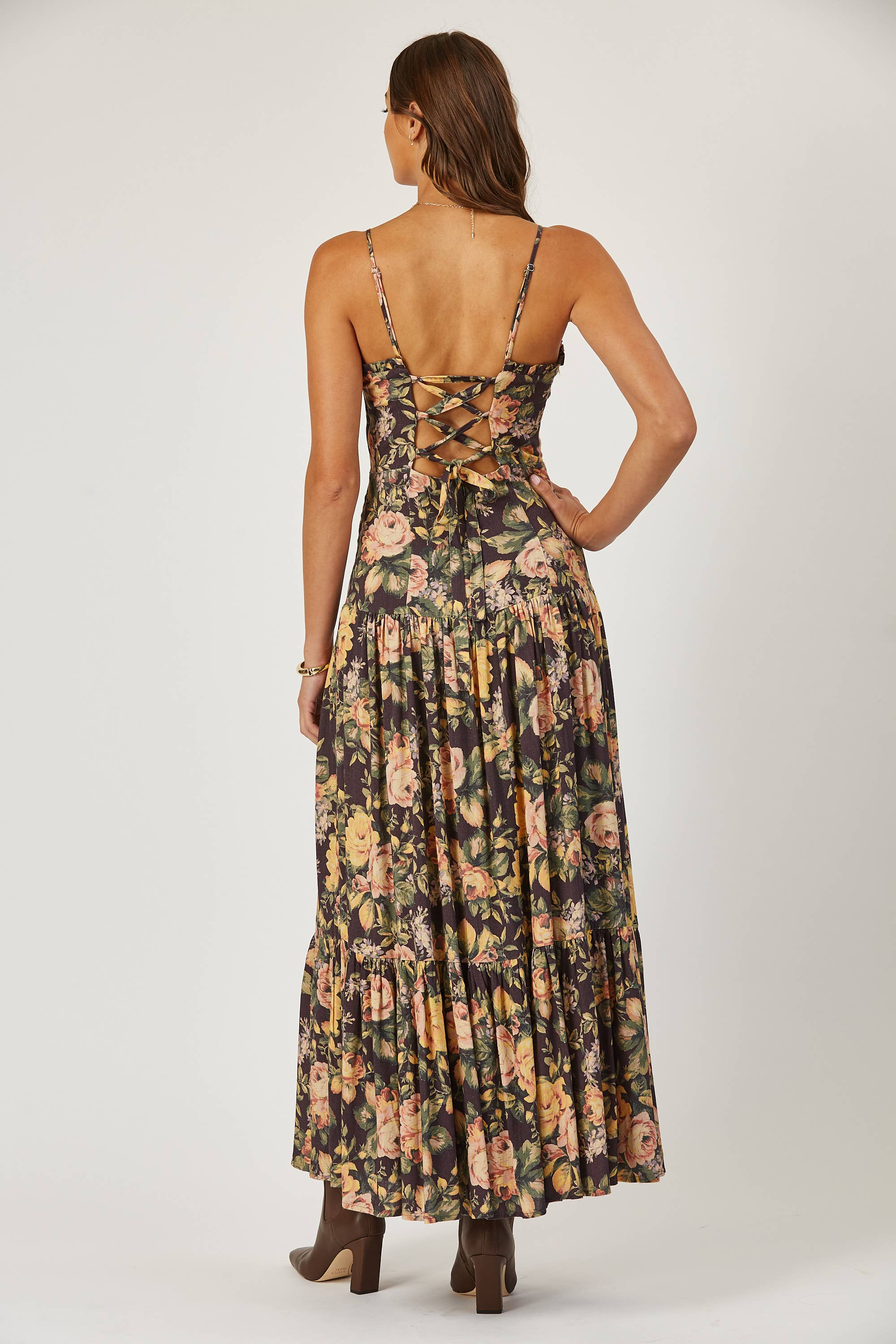 A woman with brown hair and heeled brown boots faces away, modeling the Isla Maude Priscilla laced back floral maxi dress, featuring crisscross lace-up detail, effortlessly capturing summer party style against a plain background.