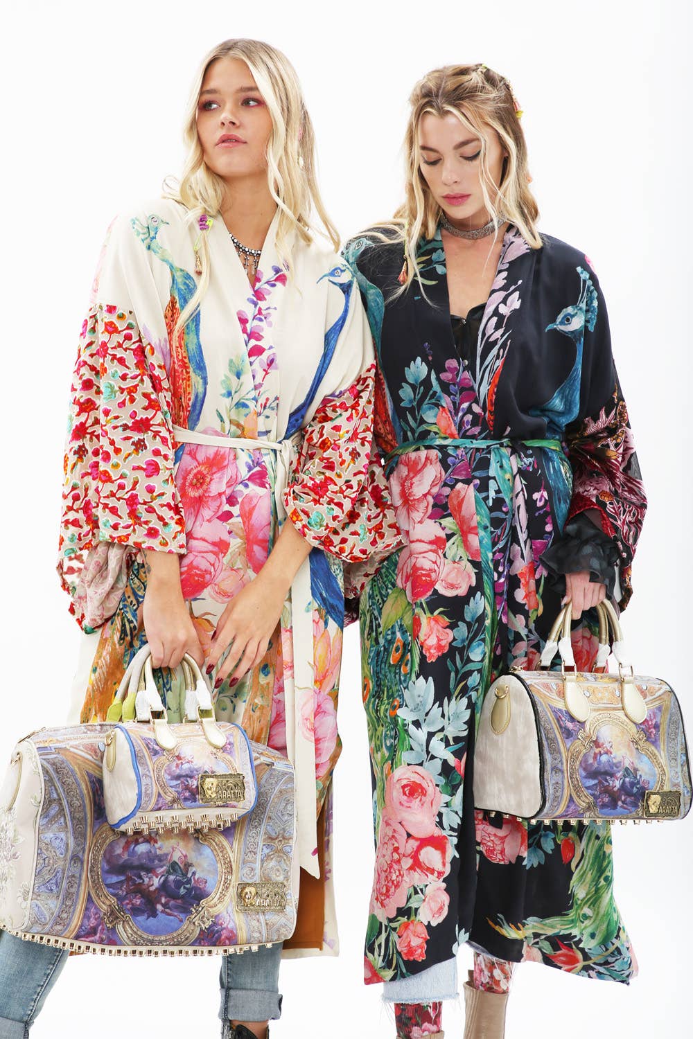 Two women with long blonde hair and confident expressions wear vibrant, floral Aratta Sweet Fantasy Kimonos—one featuring burnout velvet sleeves—and display ornate baroque-style handbags against a plain white background.
