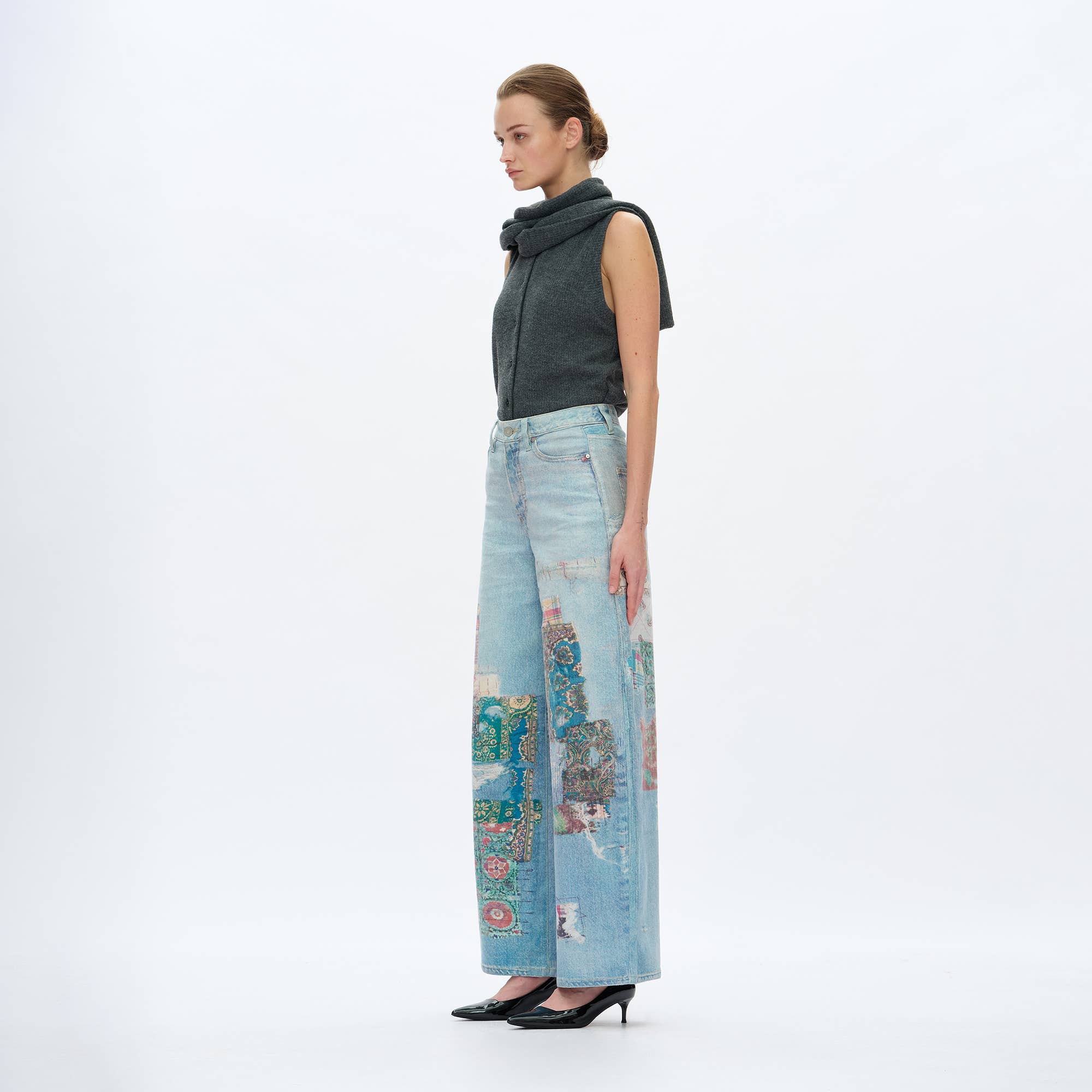 A woman stands in profile against a plain background, wearing BAYEAS Light Blue Patch Pattern High-Rise Barrel Jeans and a sleeveless gray top with a scarf-like collar, paired with black high heels.