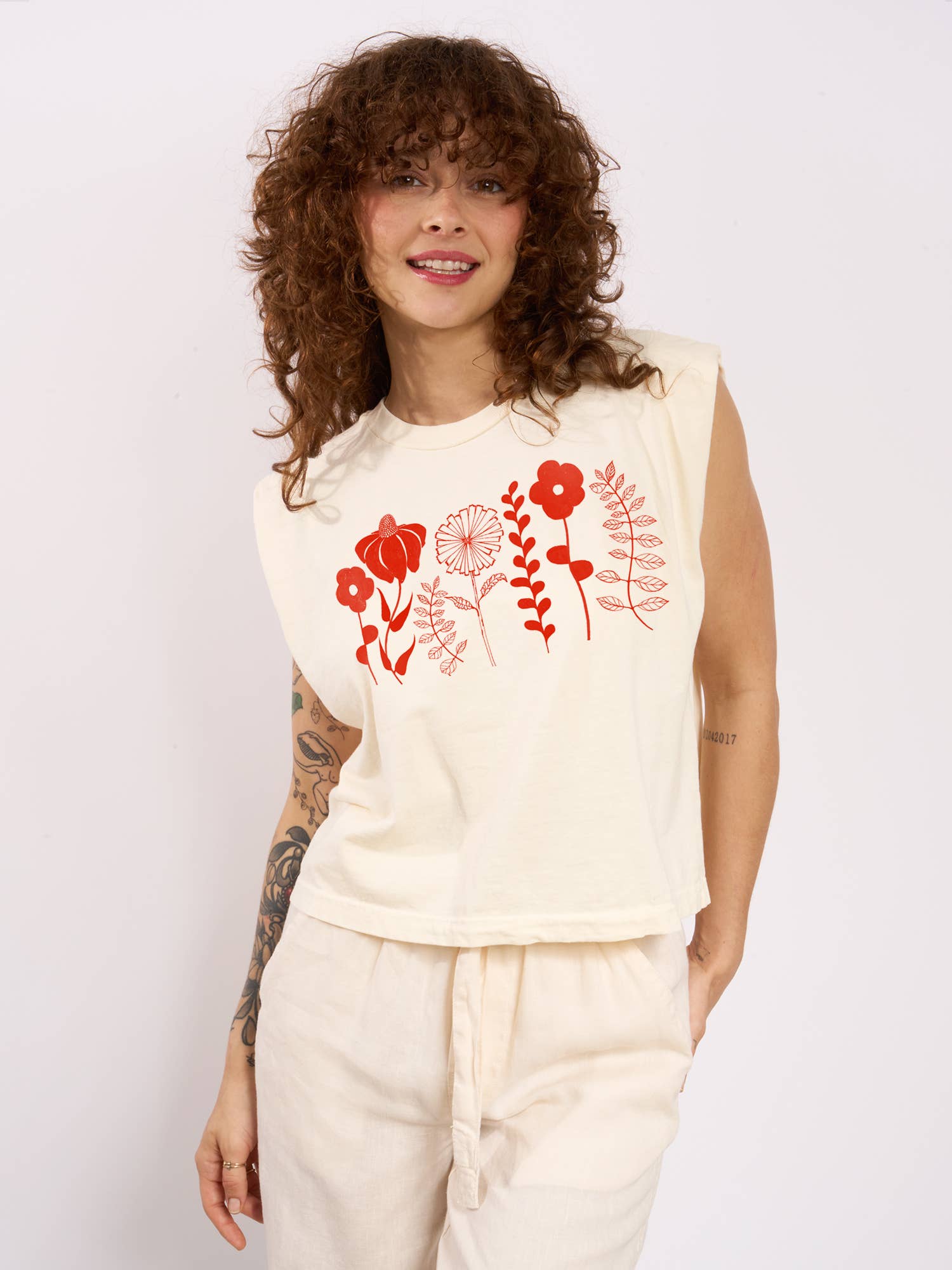 A woman with curly hair smiles, wearing a Supermaggie @ Faire Spring Flowers Maisie Heavyweight Muscle Tee with red floral designs and cream pants. She has tattoos on her arm and stands against a plain white background.