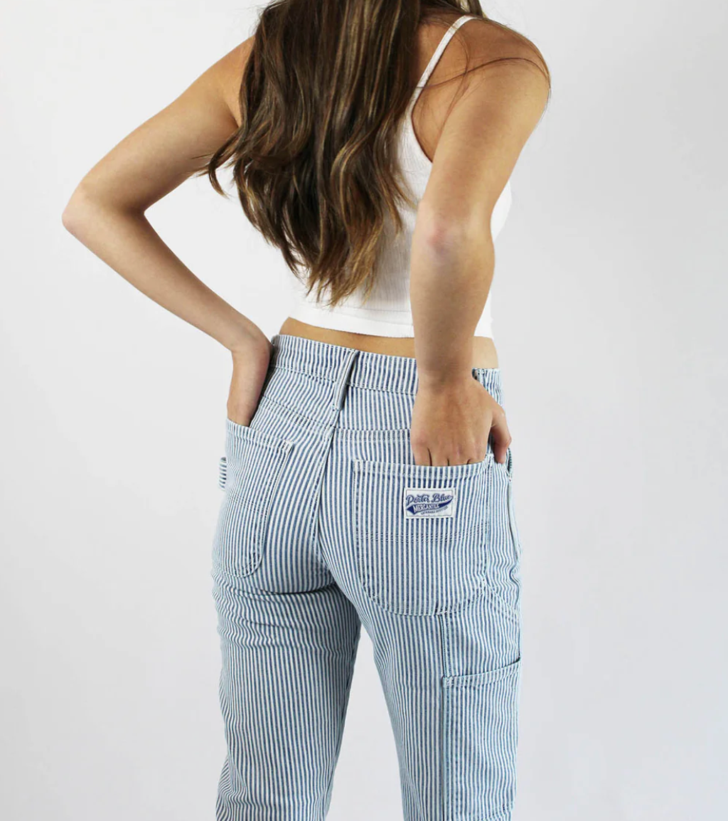 A woman with long brown hair, wearing a white crop top and Porter Blue Apparel’s PEYTON PAINTER IN PACIFIC WASH relaxed straight carpenter pants, stands with her back to the camera and her hands in her pockets.