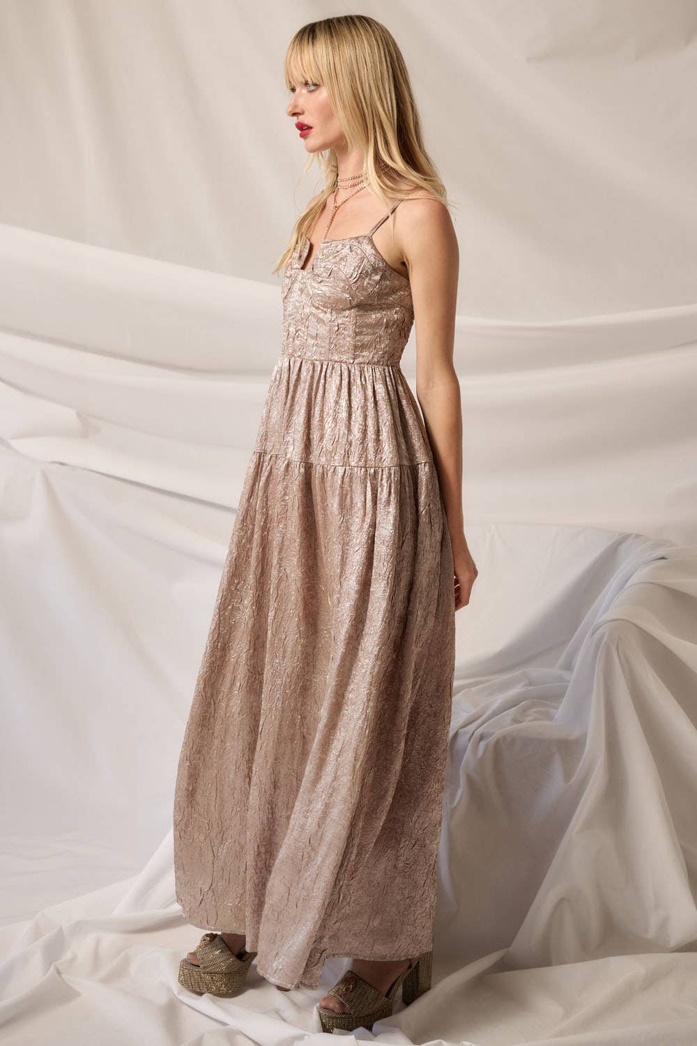 A woman with long blonde hair and red lipstick models the Promesa USA Textured Metallic Fit-and-Flare Corset Maxi Dress, standing sideways in platform shoes against draped white fabric.