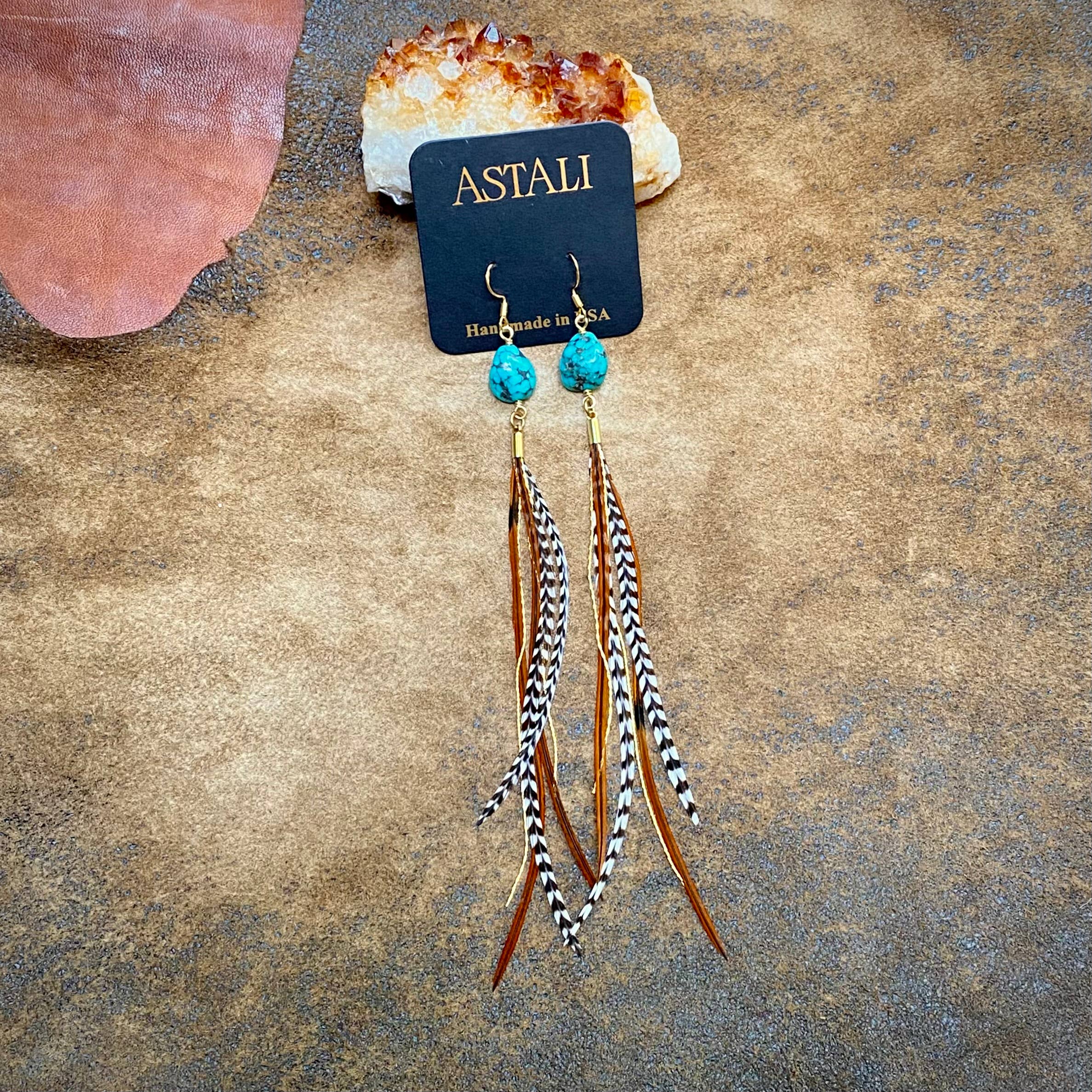 The Turquoise & Mini Feather Earrings - Grizzly & Fawn/Gold by ASTALI feature long turquoise accents with black-and-white striped and brown feathers, displayed on a branded card. These mini feather earrings add a stylish touch to any look.