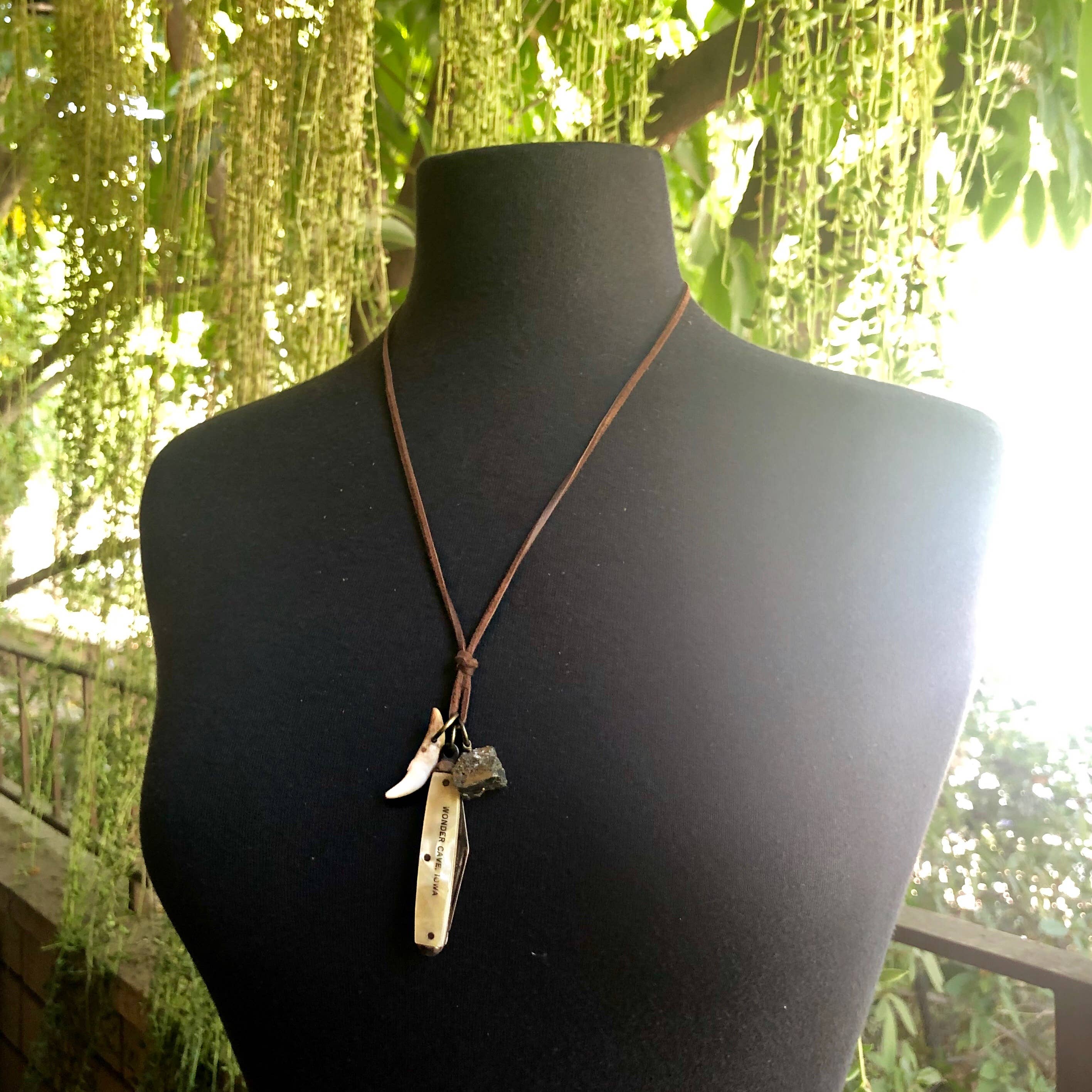A black mannequin bust displays ASTALIs Vintage Pocket Knife & Coyote Tooth Necklace—featuring a pyrite nugget, coyote tooth, and vintage pocket knife pendant on deerskin leather—set against a lush green outdoor background.