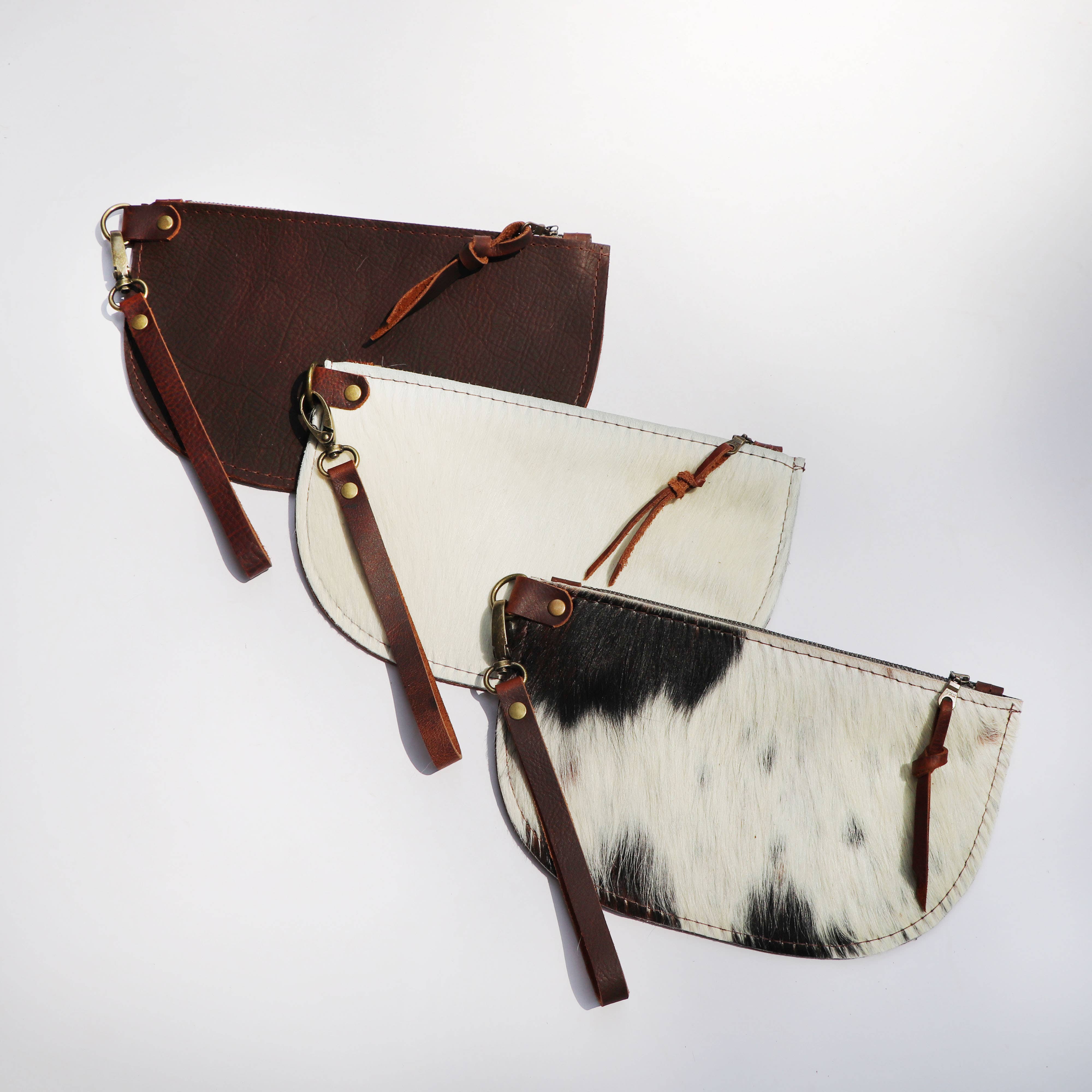 Three Hawks & Doves Large Half Moon Cowhide and Leather Zip Pouches with Wristlet are arranged diagonally: brown cowhide on top, white with brown accents in the middle, and a black-spotted pouch at the bottom. Each has a detachable wristlet strap and metal clasp.