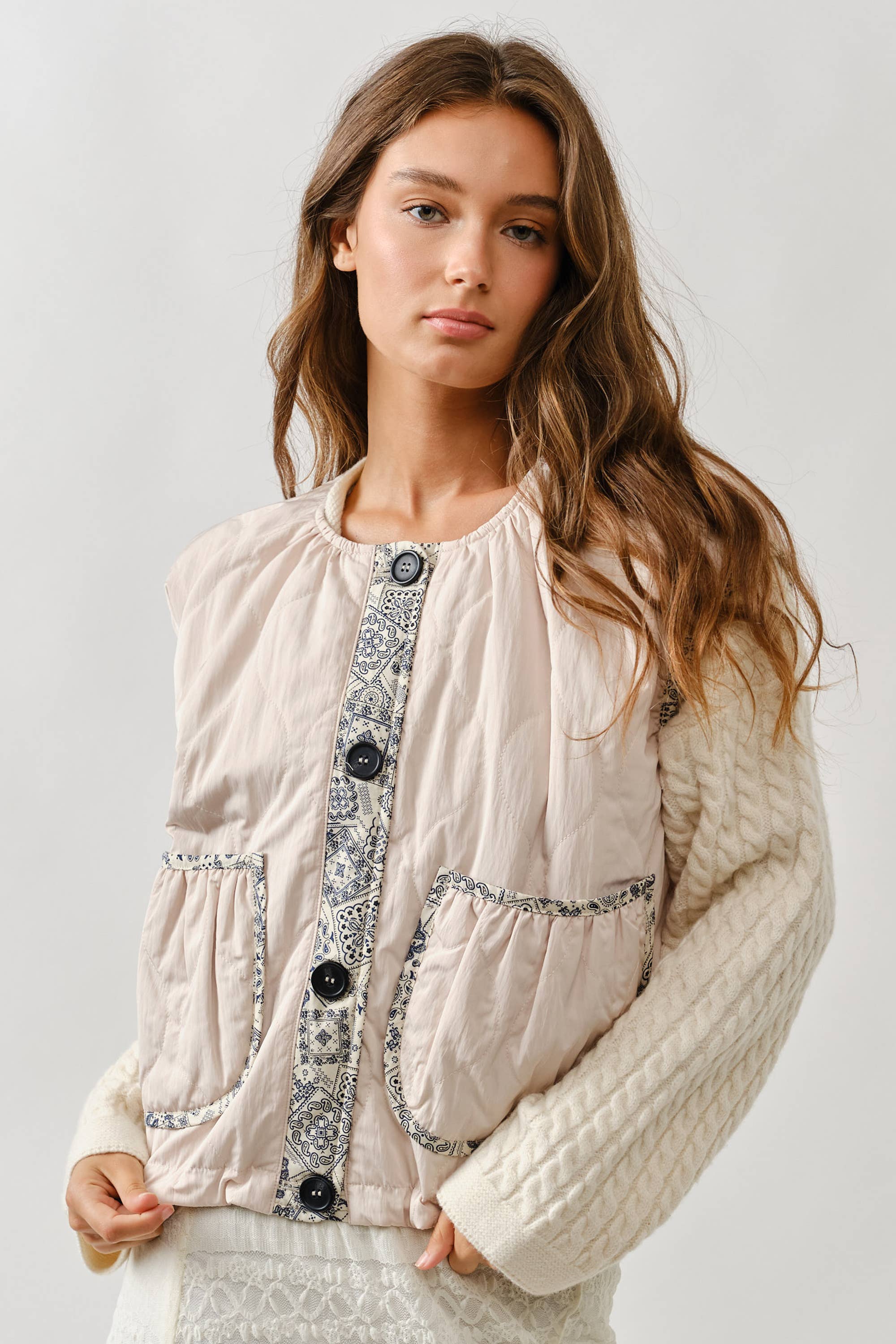 A woman with long brown hair wears the Aaron & Amber Contrast Detail Lightweight Vest—a layering essential with patterned trim, large buttons, and two front pockets—over a cream cable-knit sweater, standing against a plain light background.