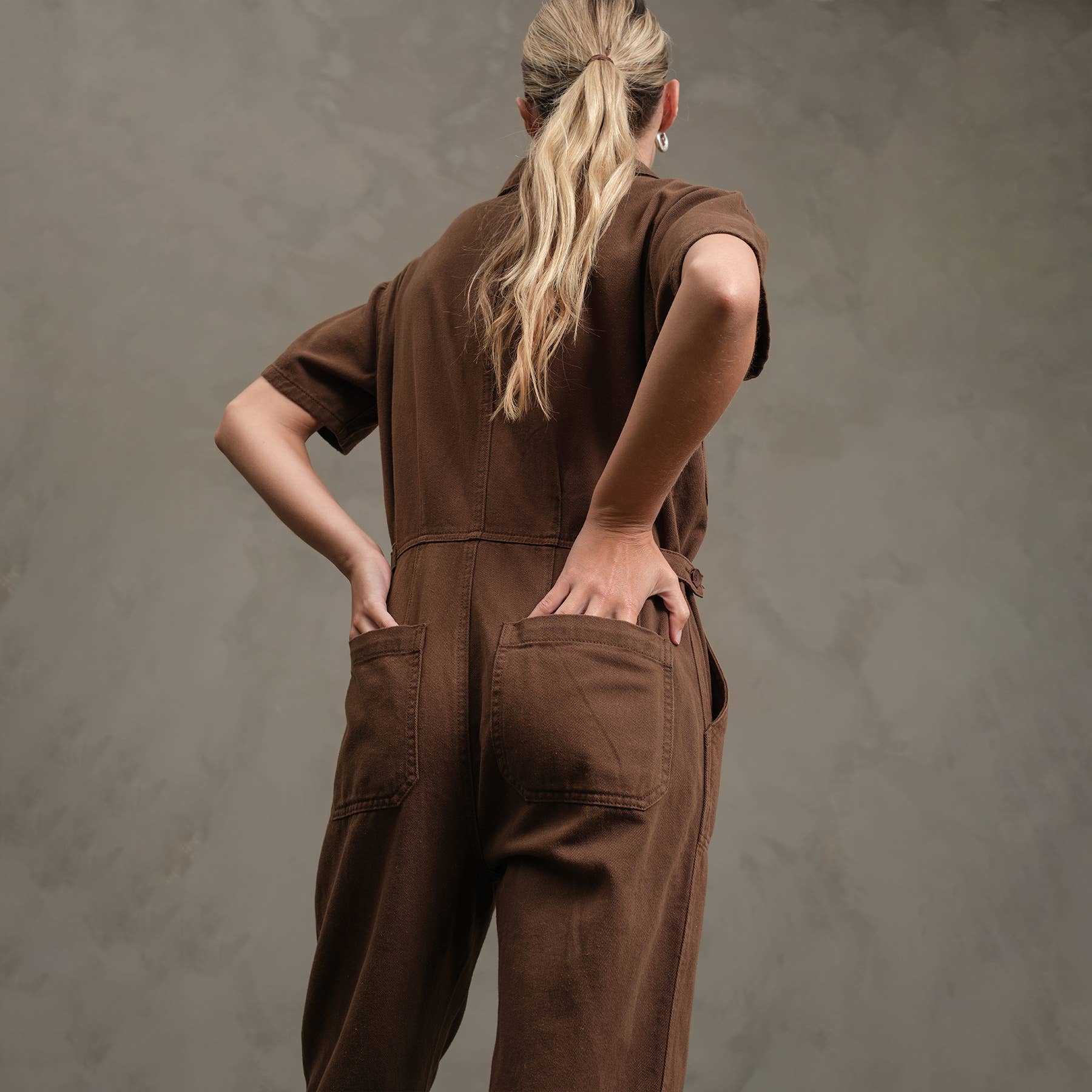 A person with long blonde hair in a ponytail stands against a gray textured background, wearing the RAISONNEL Field Jumpsuit (RJP1001) with short sleeves and both hands in their back pockets.