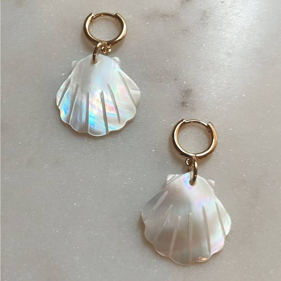 The Scallop Hoop Earrings by Tramps+Thieves feature two gold-filled hoops, each with a dangling iridescent shell charm, for a coastal-inspired look displayed on a light marbled surface.
