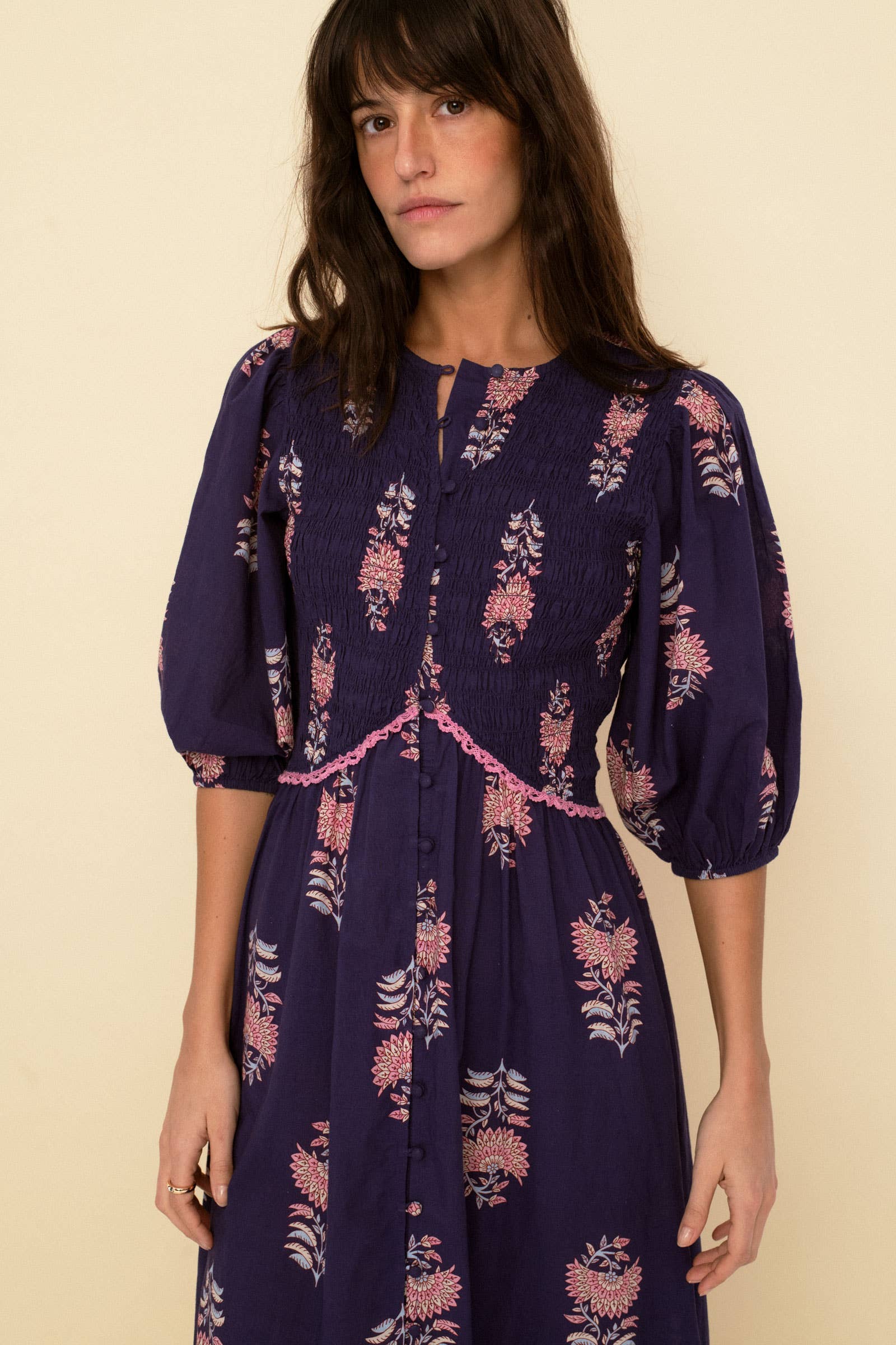 A woman with long brown hair stands against a beige background wearing the Magnolia dress blue by Sonmer Shop USA, a romantic vintage piece featuring puffed three-quarter sleeves and a pink floral pattern.
