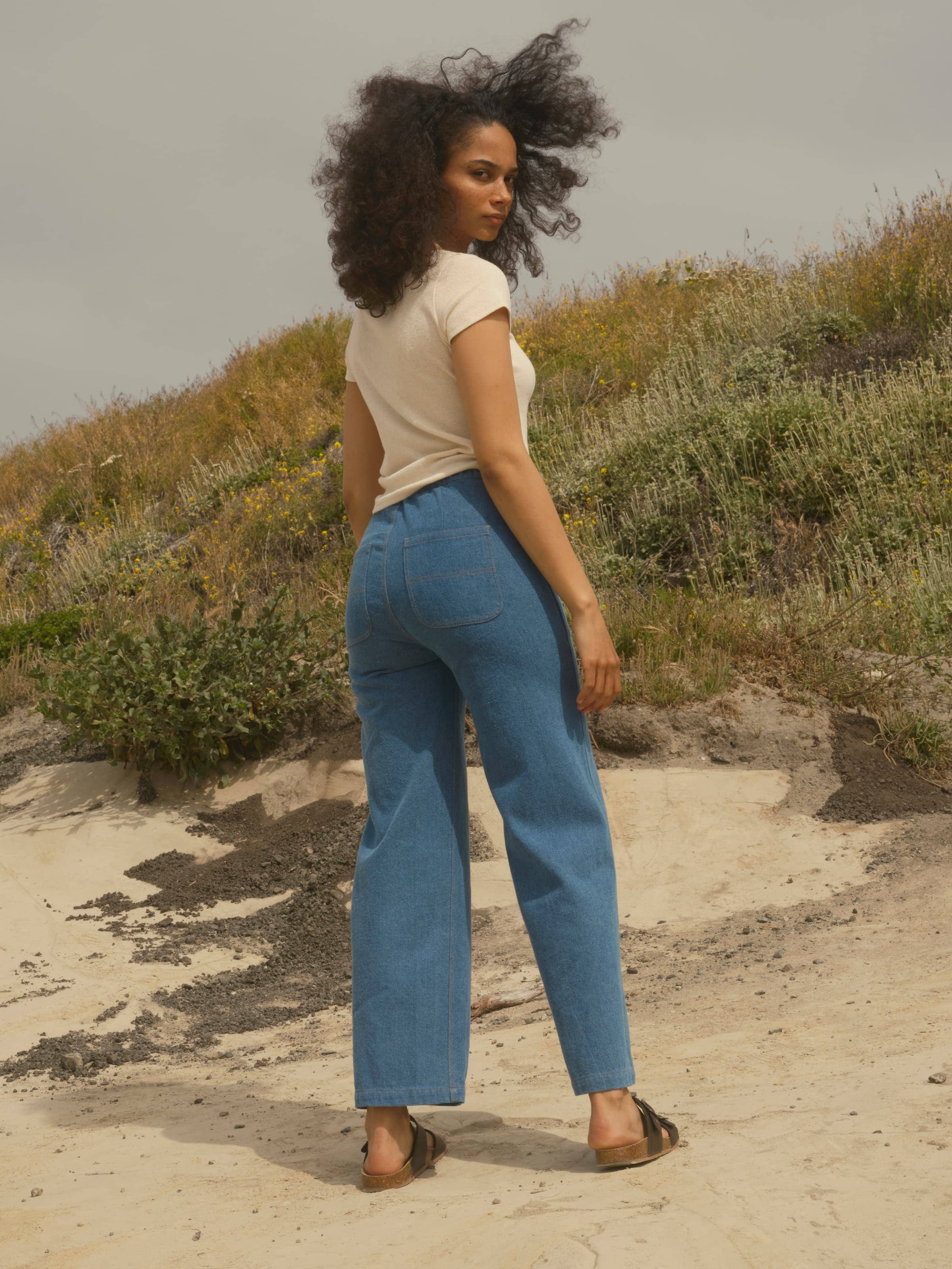 A woman with curly hair stands outdoors on sandy ground, wearing a short-sleeve beige top, Mollusk Painter Pants in Indigo (100% cotton denim), and sandals. She looks over her shoulder at grassy hills beneath a cloudy sky.