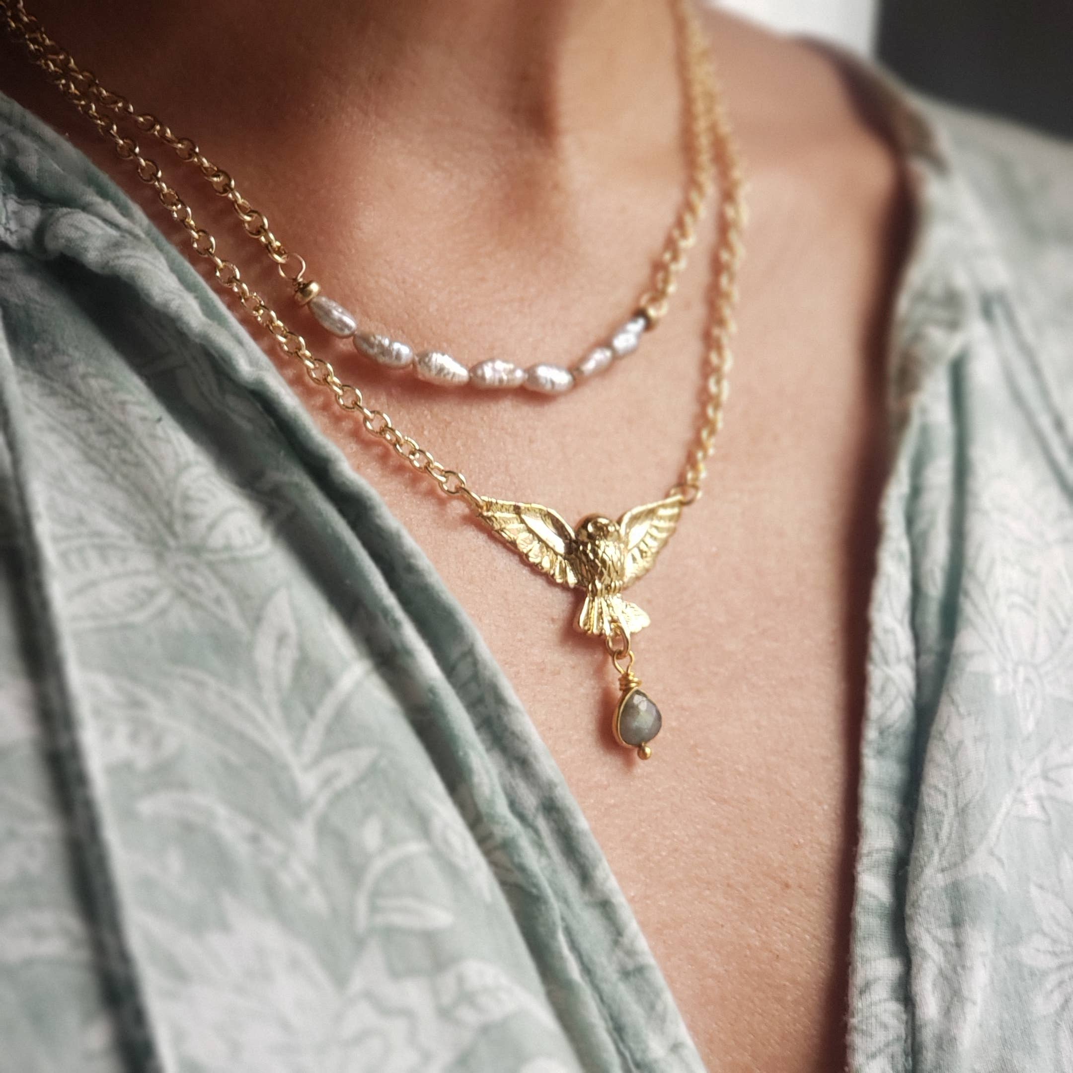 A close-up shows a person in a light green patterned outfit wearing two necklaces: one strung with small white pearls, and the Golden Owl of Intuition Labradorite Pendant Necklace by Dreamers of Dreams.
