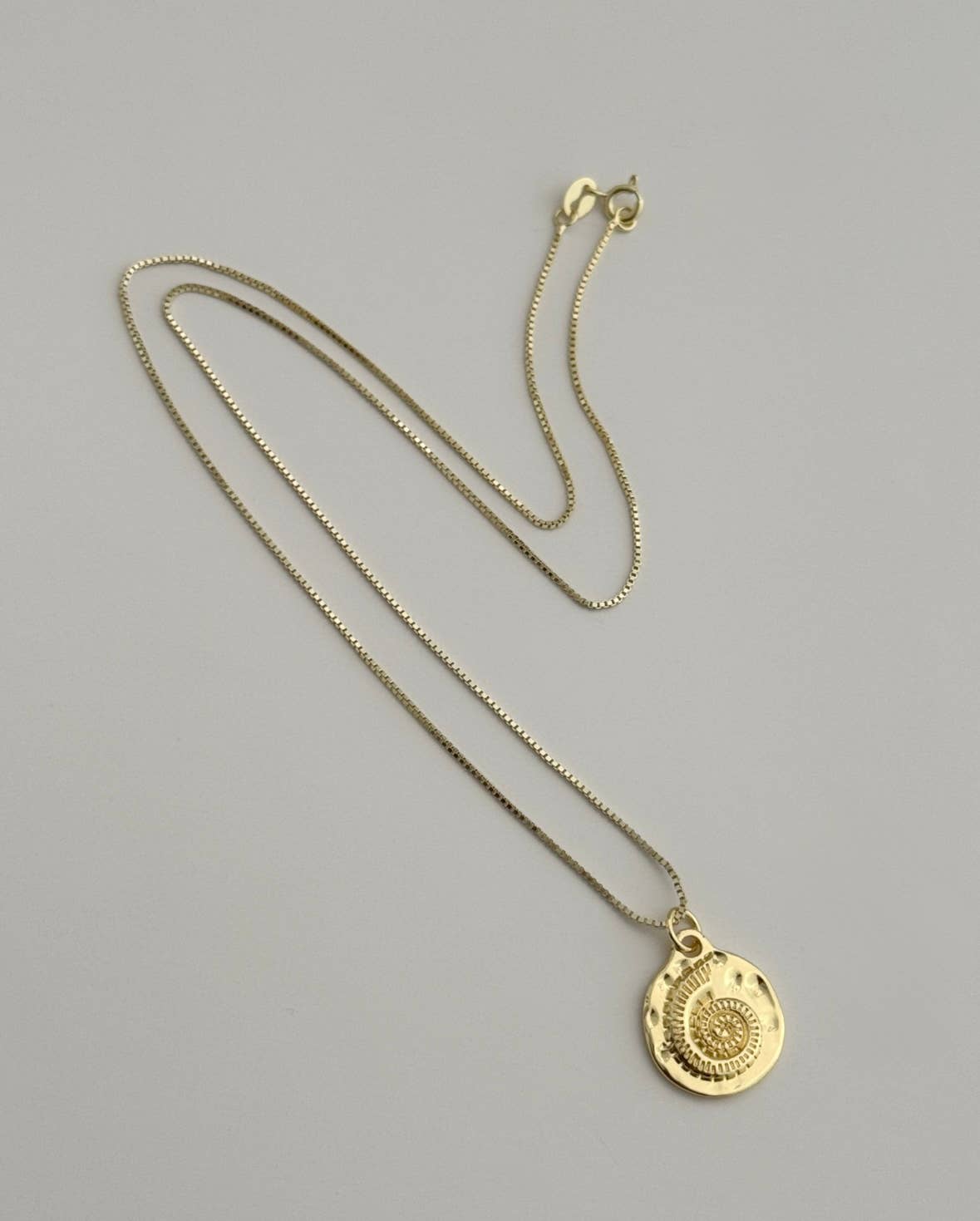The Mar Medallion Necklace by Tramps+Thieves features a gold vermeil thin chain and a round, textured shell pendant with a concentric spiral design, shown against a light gray background.