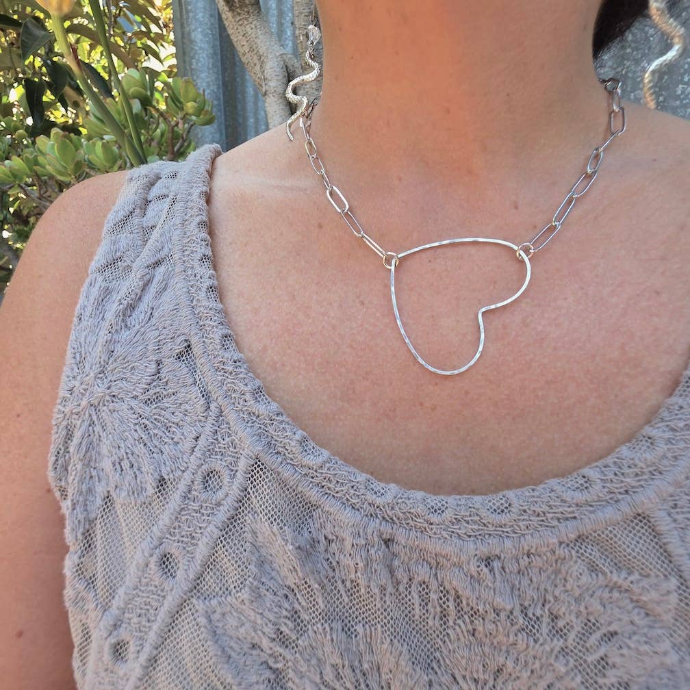 A person wears a textured, sleeveless grey top and the Dreamers of Dreams Big Silver Heart - Paperclip Chain Necklace in silver-plated brass, with green leaves and a metal fence in the background.