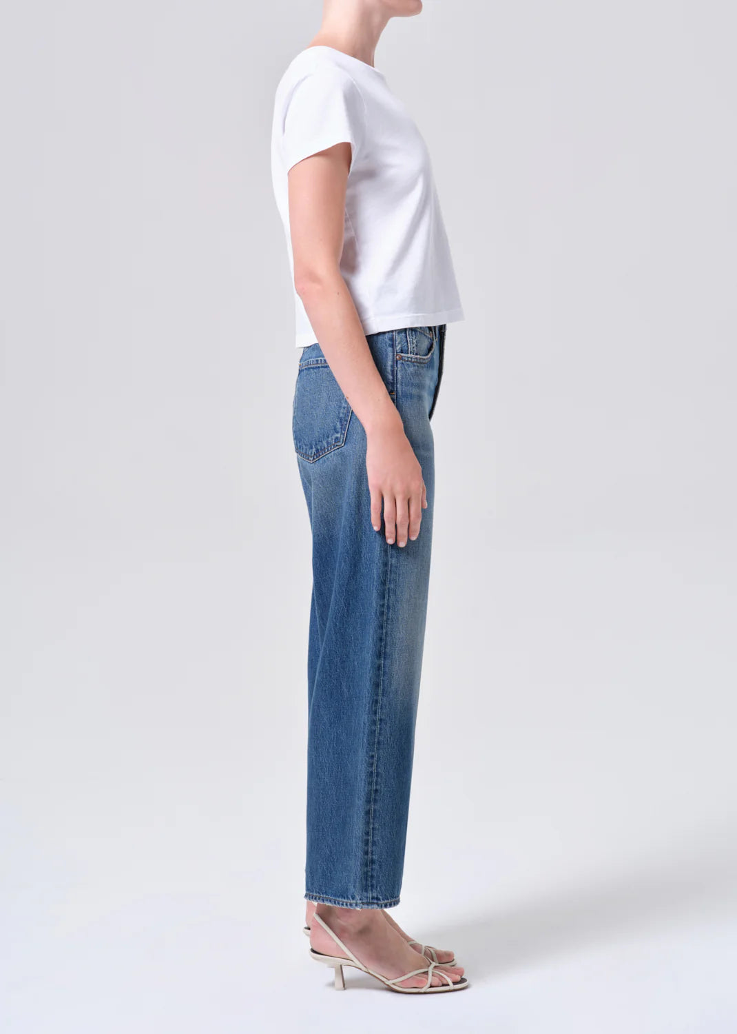 Side view of a model wearing REN JEAN in chapter blue denim jeans with a white top and strappy heels