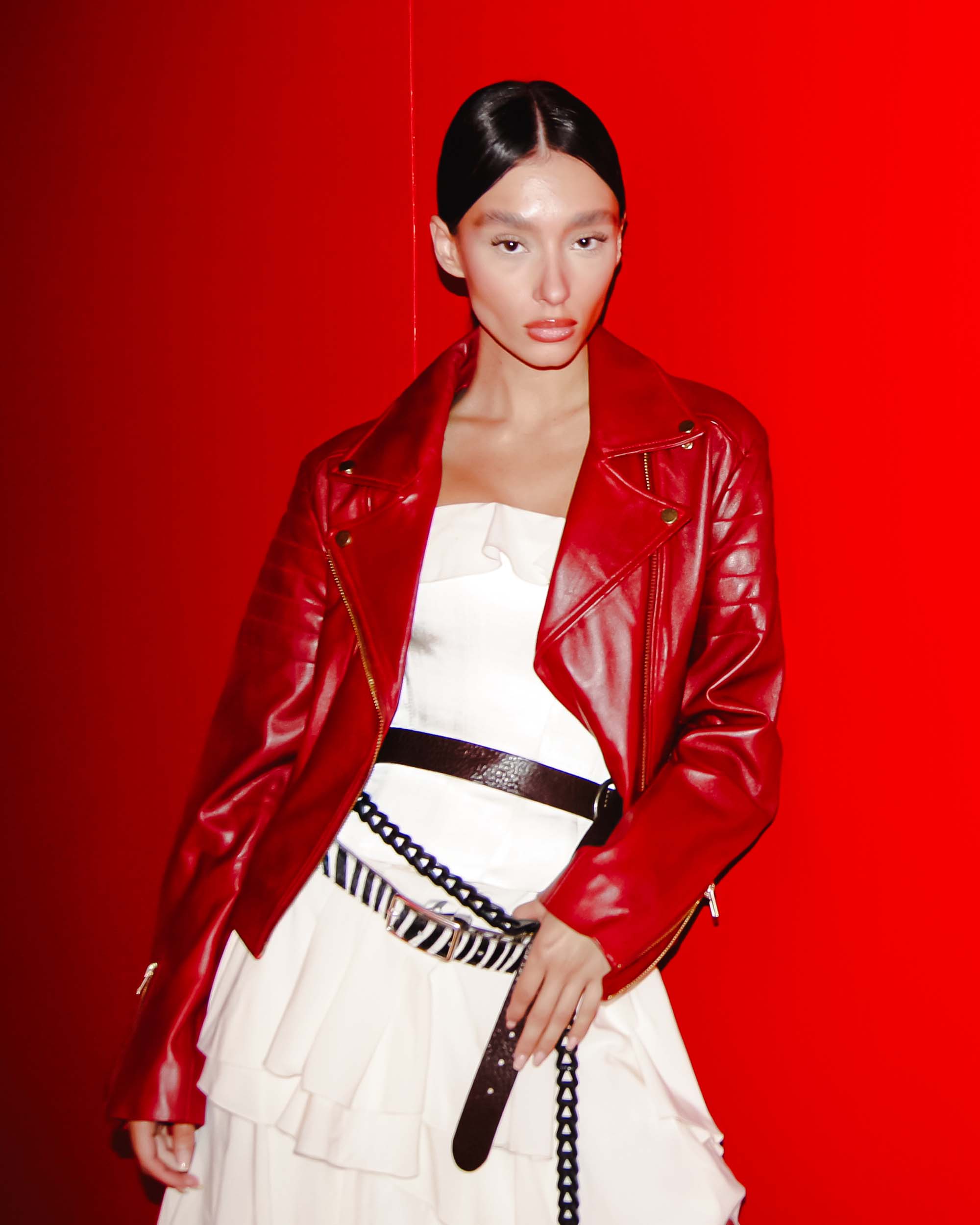 Model wearing a bold red Eureka | Leather Biker Jacket over a white dress with layered details and belts.