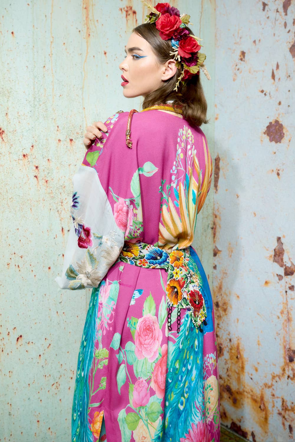 A woman in Aratta’s Marie Antoinette Kimono—with embroidered floral sleeves—stands in profile by a rusty wall. Red flowers and gold leaves accent her hair, while bold makeup completes this striking statement look.