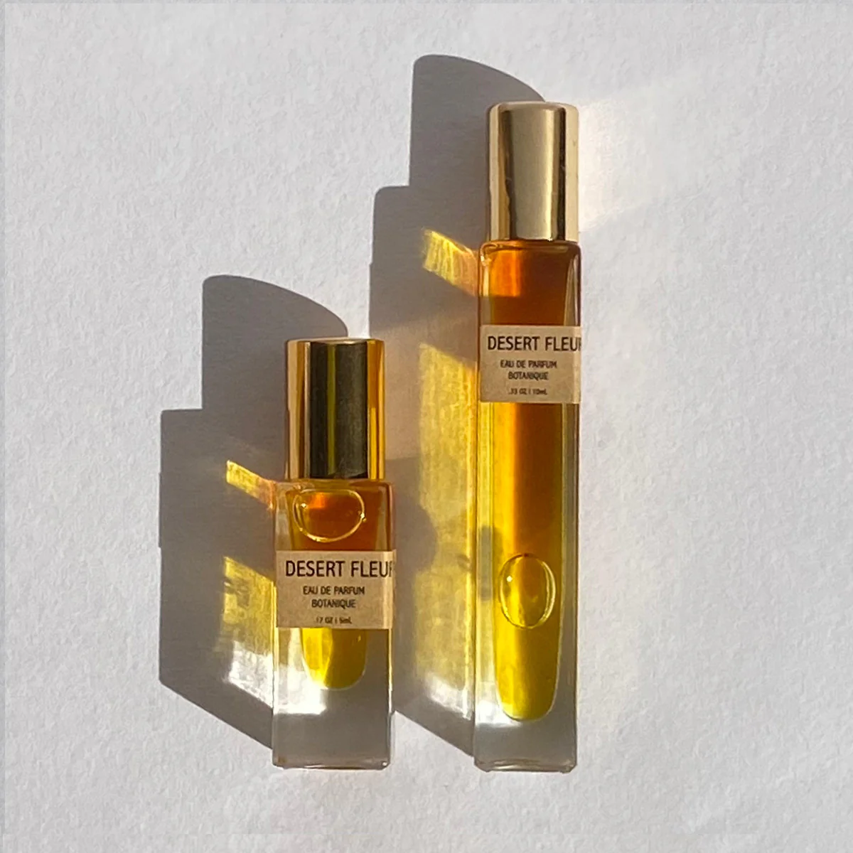 Two perfume bottles with golden caps casting shadows, labeled Desert Fleur Botanical Perfume Rollerball 10mL | Earthy & Iconic