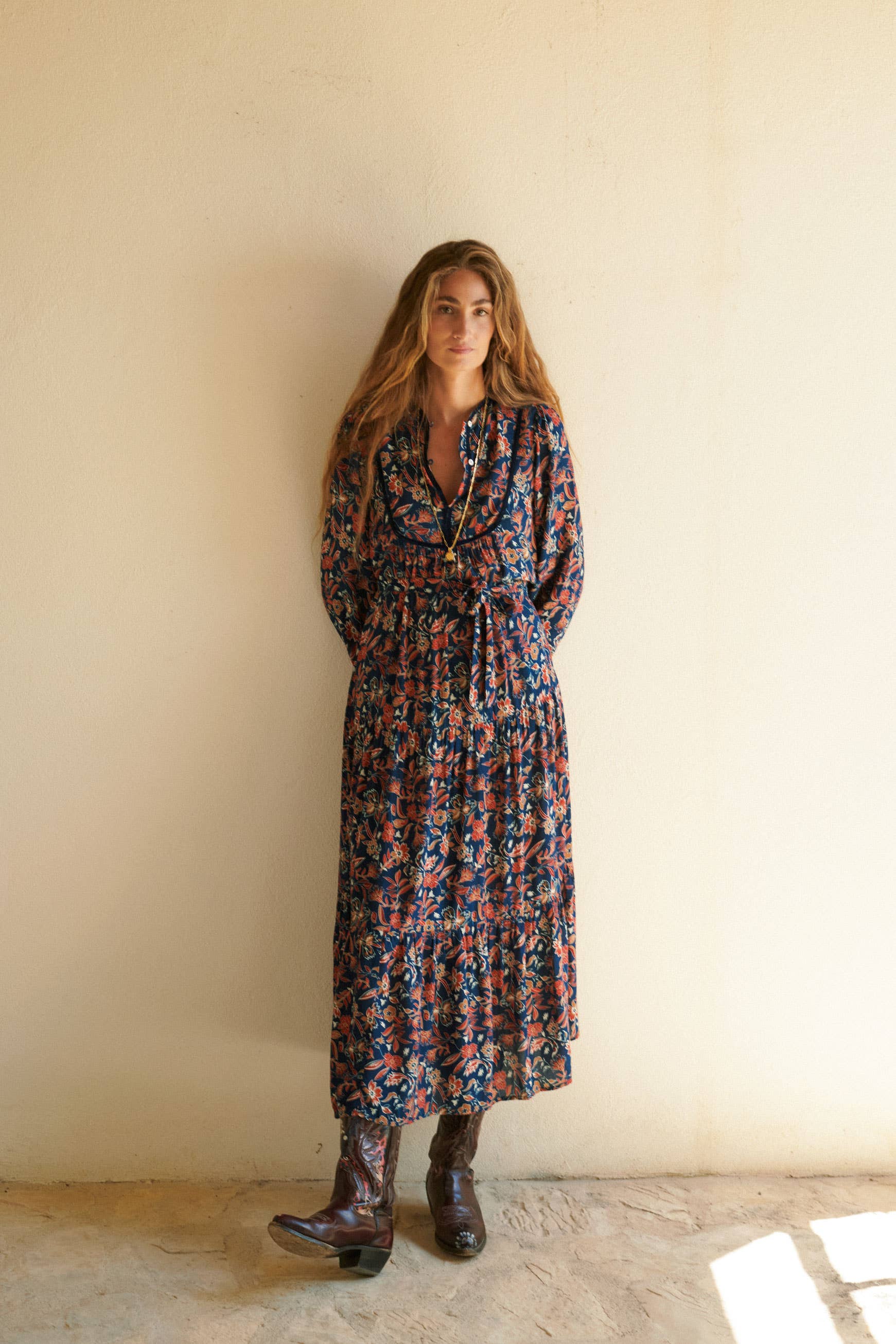 A woman with long brown hair stands against a beige wall, wearing the ankle-length Aura Dress Navy from Sonmer Shop USA. The bohemian maxi dress features long sleeves and an adjustable V-neckline, giving her look a relaxed charm.