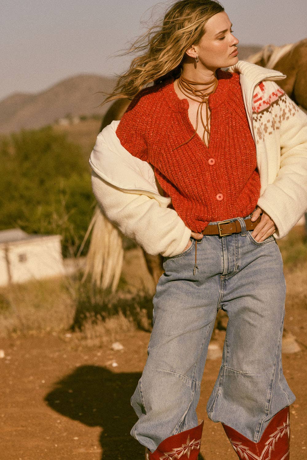 A woman outdoors wears the Promesa USA Confetti Button-Front Cardigan Sweater, an off-white patterned jacket, loose-fit jeans, and red cowboy boots. Her windswept hair and a glimpse of a horse enhance the rural, hilly landscape.