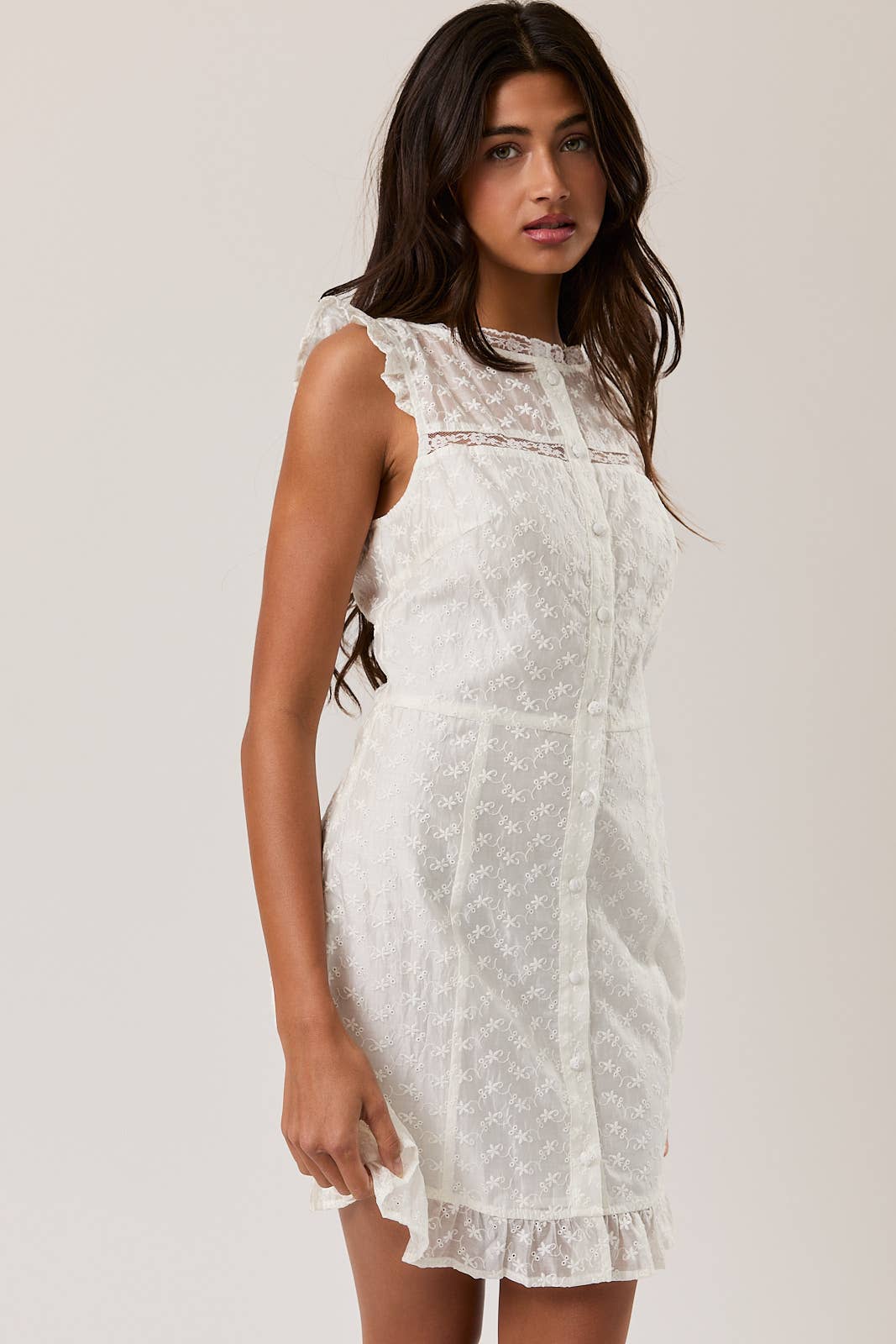 Woman wearing a white Eyelet Lace Mini Dress with ruffle details and delicate floral embroidery standing against a plain background