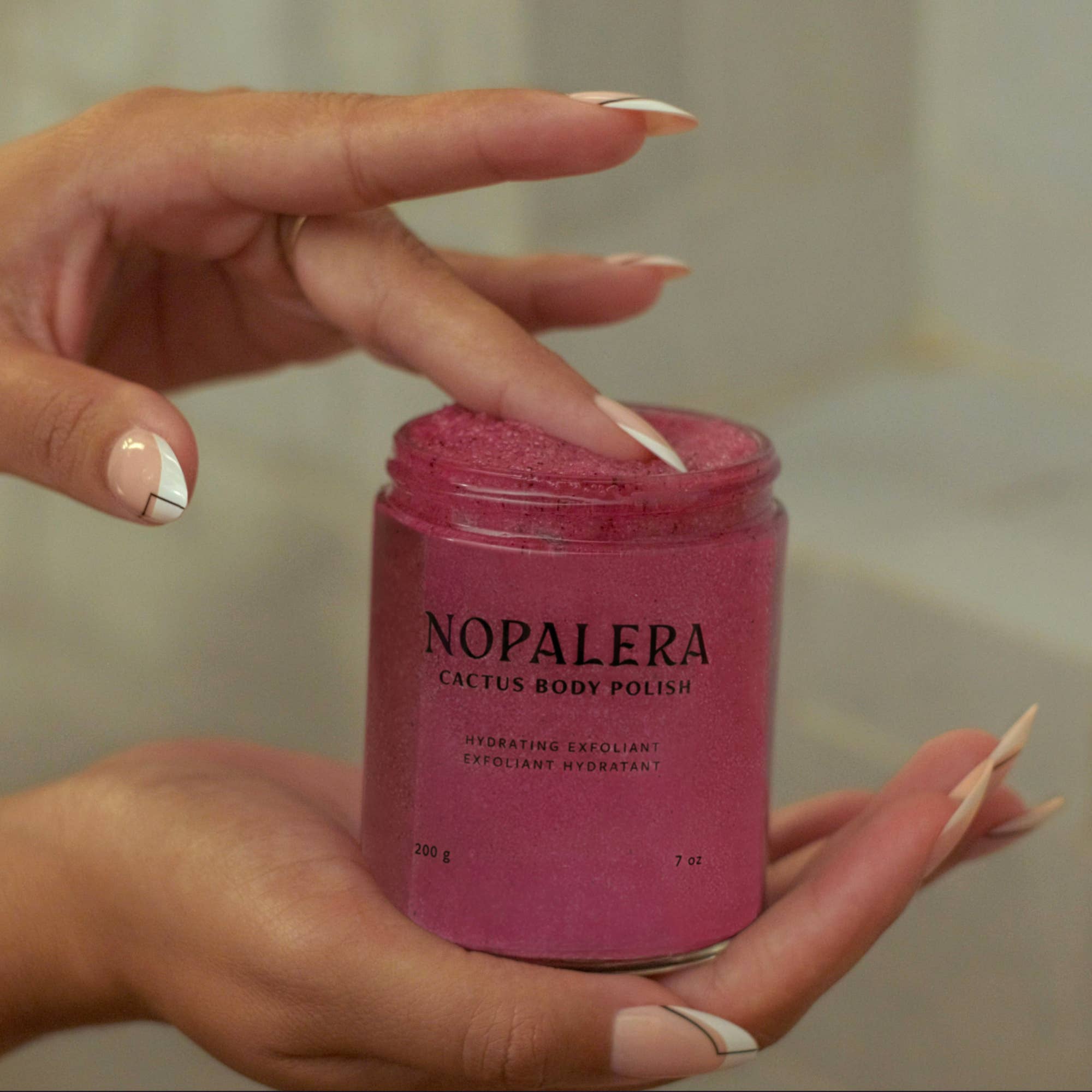 Hand dipping finger into vibrant Hibiscus Cactus Body Polish jar held by manicured, manicured hands with artistic nails.
