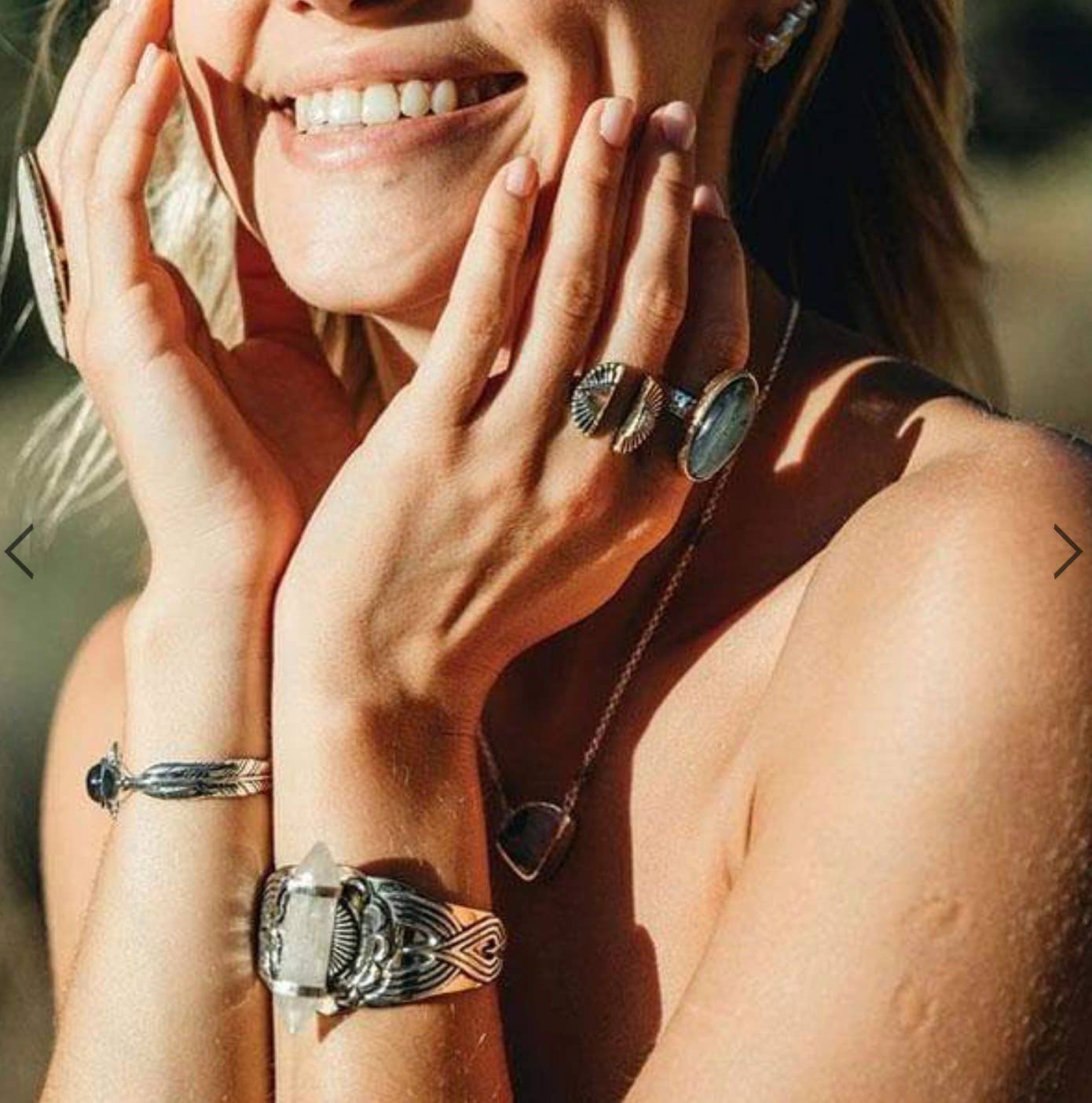 A smiling woman touches her face, sunlight highlighting her silver bahgsu jewels Shape Shift Ring and matching gemstone and feather bracelets and necklaces.