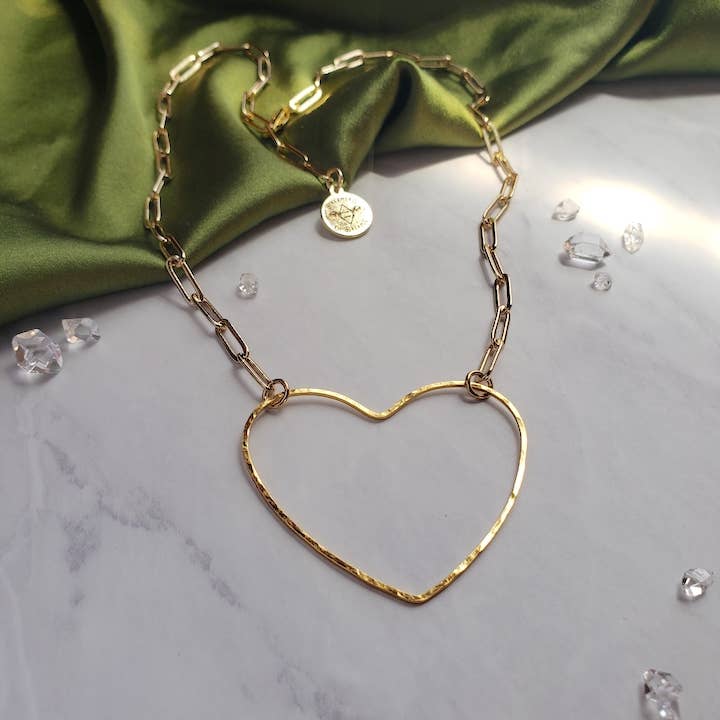 The Big Gold Heart | Paperclip Chain Necklace by Dreamers of Dreams, featuring a large open heart pendant and round charm, rests on white marble with green fabric and scattered clear gems in the background.