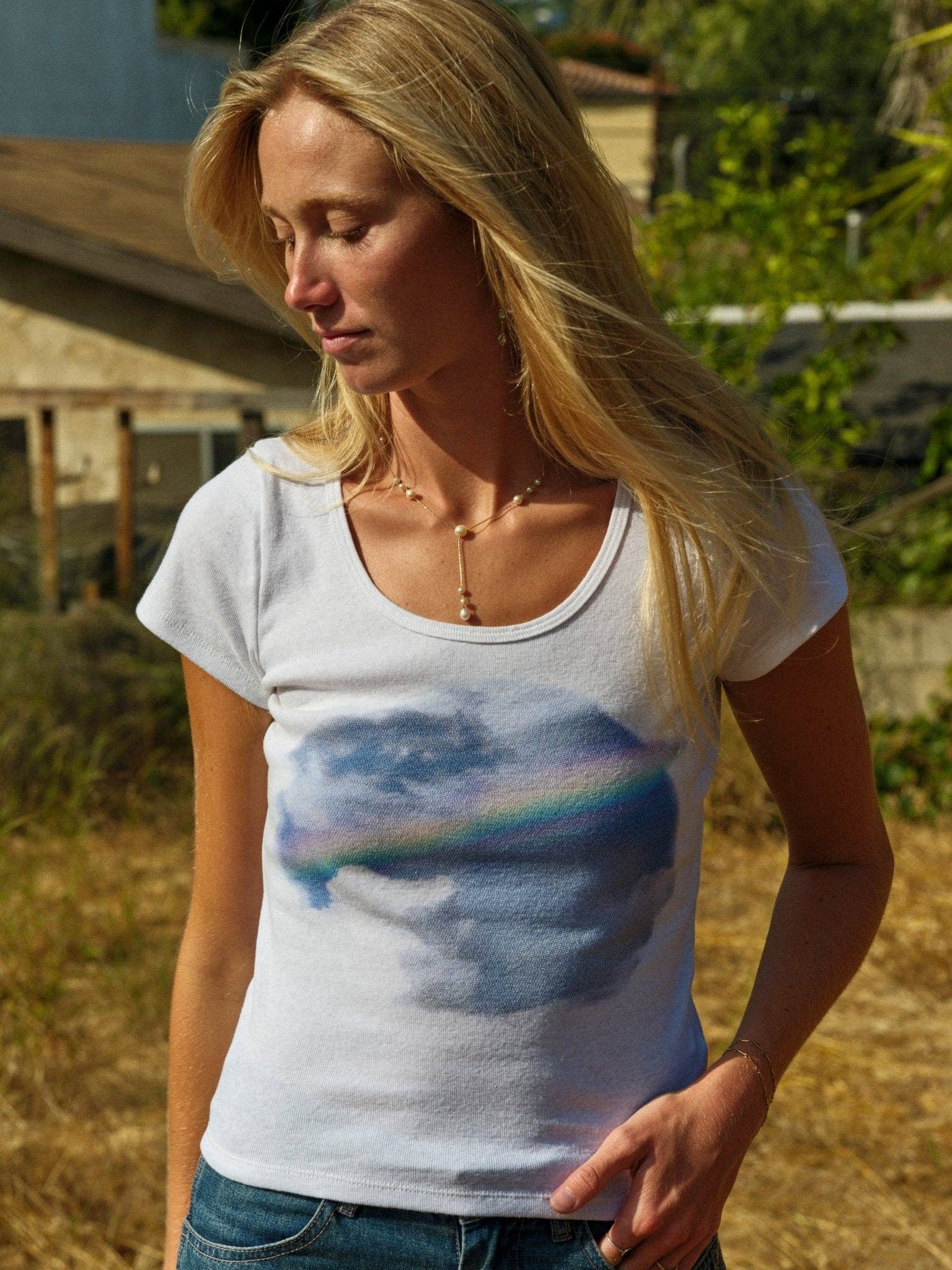 A woman with long blonde hair stands outdoors, hand in pocket, wearing the Mollusk CloudBreak Tee—crafted from sustainable hemp cotton and featuring a cloud and rainbow design. Greenery and a building appear in the background.